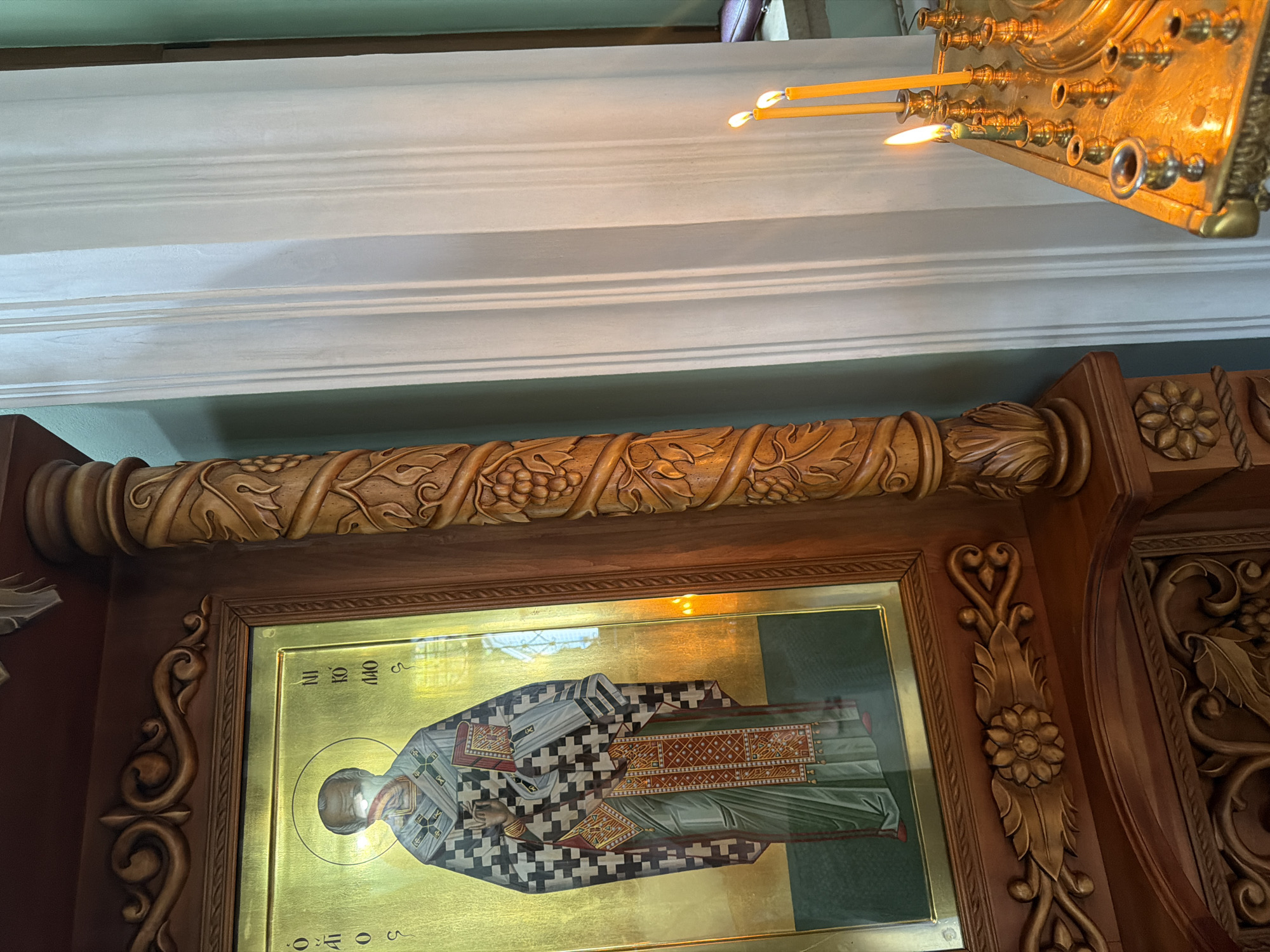 In this close-up, indoor shot, an Orthodox icon of a saint with a halo is displayed on a wooden stand. The saint is depicted wearing religious vestments and holding a book. To the right of the icon, a carved wooden column features a vine motif. Further to the right, a brass candelabrum holds several lit candles, casting a warm glow on the scene. The background consists of light blue walls and sheer white curtains. This image was captured in the city of Sochi, Russia.
