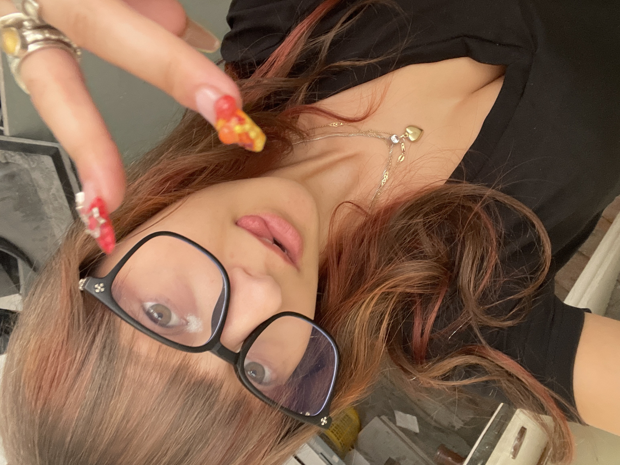 A close-up, overhead selfie taken in Raanana, Israel, shows a woman with long, reddish-brown hair, styled with pink highlights. She is wearing black-rimmed glasses and a black V-neck top. Her right hand, adorned with elaborate red and yellow nails and rings, is raised towards the camera, with her index finger extended in a peace sign. A delicate gold necklace with a heart-shaped pendant rests on her décolletage. Her tongue is visible between her lips, and her eyes are looking slightly to the side. The background is blurred, but hints of furniture and a grey wall are discernible.