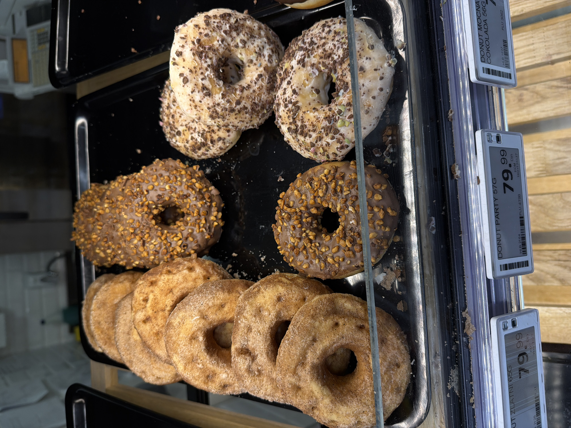 This is a close-up photograph of a display case containing various donuts, likely in a bakery or grocery store. The donuts are arranged on a black tray. The top row features two donuts covered with a white frosting and sprinkled with chocolate shavings. The middle row has two chocolate frosted donuts covered in chopped nuts. The bottom row contains approximately six to seven cinnamon sugar donuts. Adjacent to the tray, several price tags are visible. The price tag for "Donut Party 57G" is 79.99. Based on the language and price tags, it can be assumed this image was taken in Serbia, likely in the city of Vracar Urban Municipality.