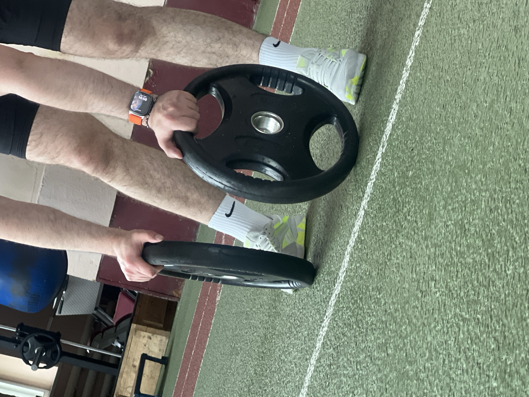 The image depicts an indoor sports facility in Poltava, Ukraine. A male individual is partially visible from the mid-thighs to the forearms, standing on a green rubberized floor marked with white and red lines. He wears black athletic shorts, white crew-cut socks with black Nike swoosh logos, and light grey athletic shoes with prominent neon yellow accents. An orange-strapped Apple Watch Ultra is visible on his left wrist, displaying a digital time of "16:59". He holds two black Olympic-style weight plates, each with a silver central hub and three integrated handles, positioned vertically on the floor, one in each hand. The plates appear to be standard sized, likely 5kg or 10kg based on the diameter. In the background, various gym equipment is discernible, including a suspended blue punching bag, a weight machine with a black plate, a maroon upholstered exercise bench, and wooden and blue step platforms. The walls behind are two-toned, with a lighter upper section and a darker lower section. A window is partially visible in the upper left background.