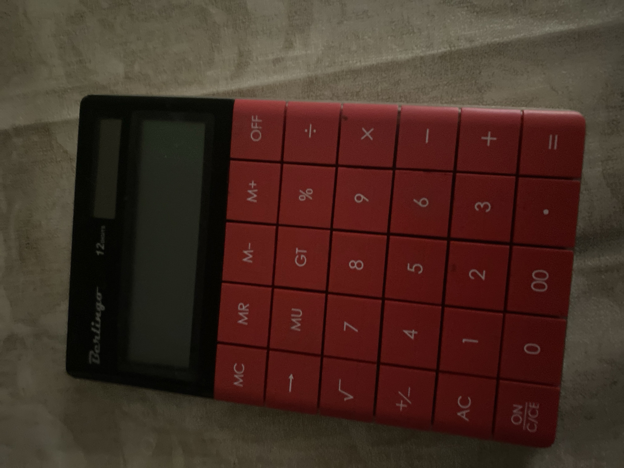 A red and black calculator is lying on a light-colored, textured surface. The calculator has a screen at the top and a keypad below. The keypad features red buttons with white text and symbols. The buttons include numbers (0-9), basic arithmetic operations (+, -, x, ÷), memory functions (MC, MR, M-, M+), a square root symbol (√), a percentage symbol (%), an "AC" (All Clear) button, a "C/CE" (Clear/Clear Entry) button, a "ON/OFF" button, and an equals sign (=). The calculator is branded "Berlingo" and indicates "12 digits". The image is taken from a slightly elevated angle, focusing on the calculator. There are no people visible, and no indications of the city of Tolyatti, Russia, or any specific urban environment.