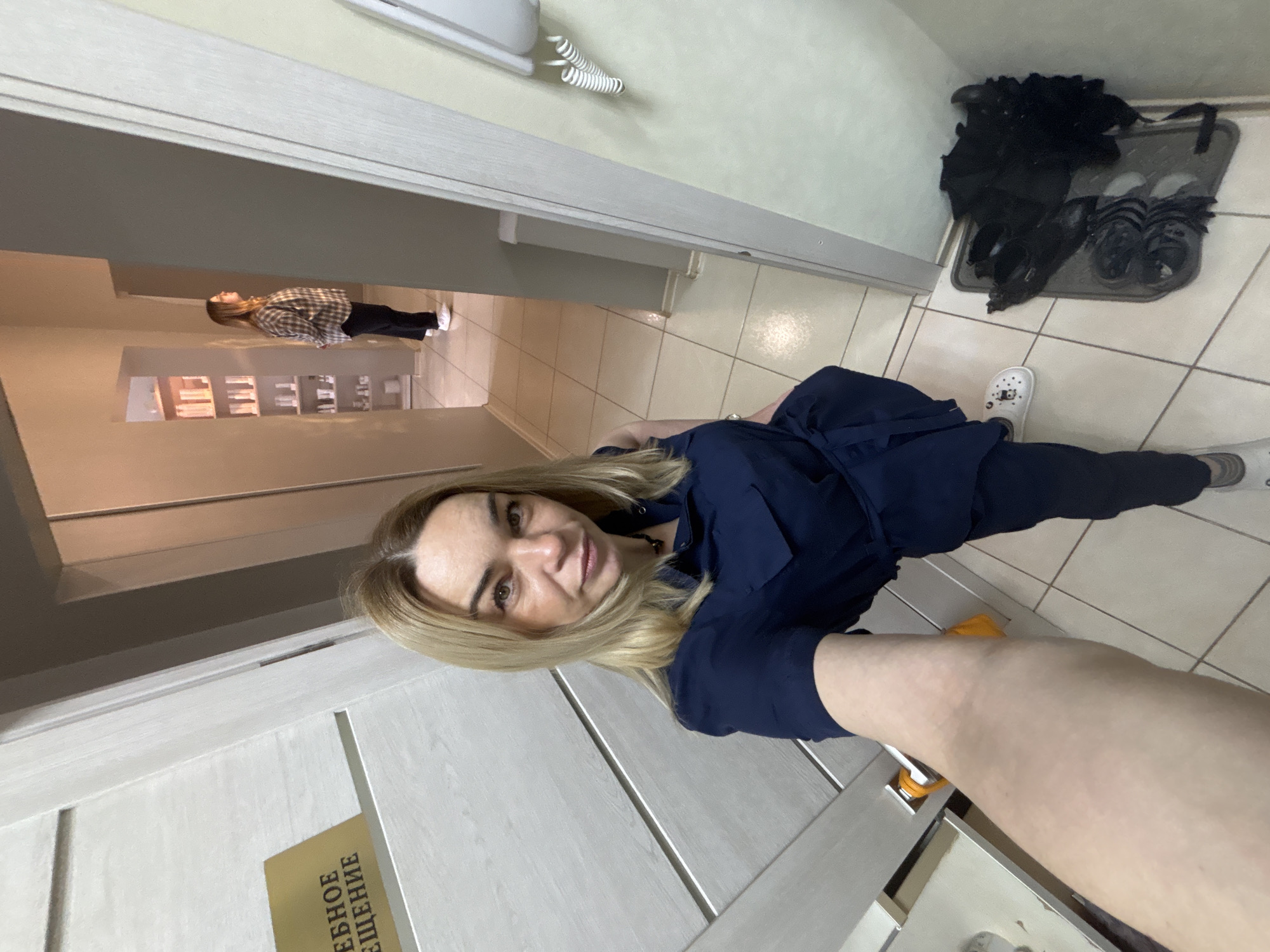 A medium shot angled down shows a woman standing in a hallway, taking a selfie in Unknown, Unknown. The woman has shoulder-length blonde hair and is wearing a navy blue dress. The hallway has tiled floors and leads to another room with shelves and a person in a plaid shirt. Near the doorway are a mat, shoes, and a black item (likely a jacket). The door to her right has a sign in Russian, indicating "Ready Solution".
