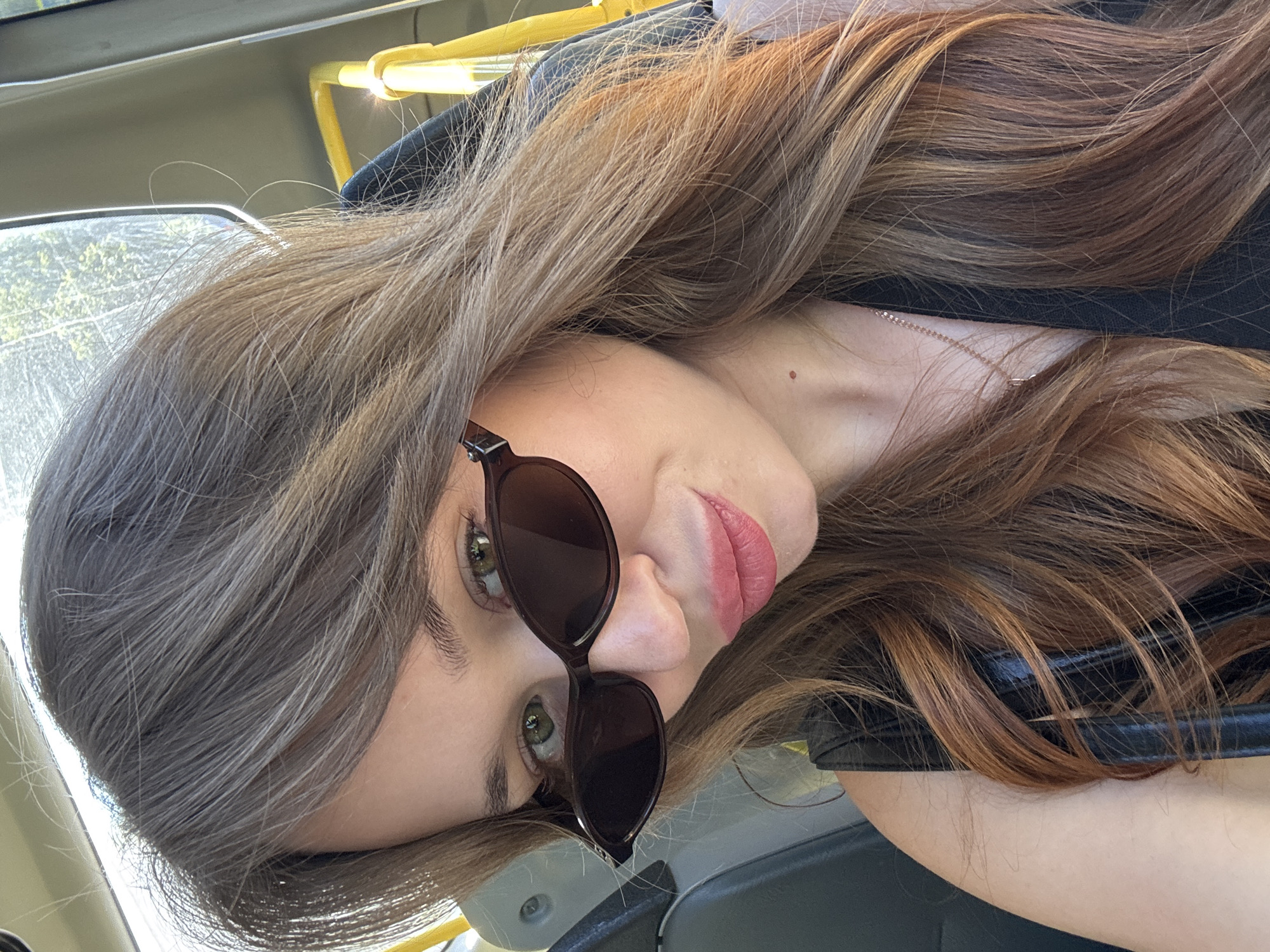 A close-up, selfie-style photograph features a young Caucasian woman with light brown hair and green eyes, wearing dark sunglasses and a black top. She is seated inside a vehicle, possibly a bus, indicated by the yellow handle and gray seat visible in the background. Sunlight streams in from a window, illuminating her hair with warm tones. She is looking towards the camera with a slight smile, and her lips are painted a rosy pink. A delicate gold necklace is visible around her neck. The background suggests an urban environment, with trees visible through the window, hinting at a location in Chaikovsky, Russia.