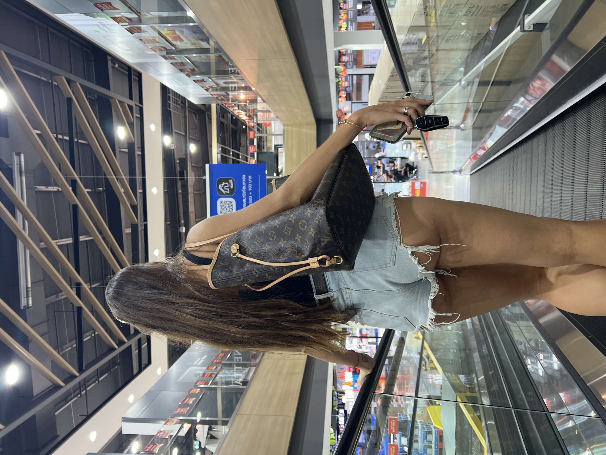 A person with long, dark hair is walking up an escalator in a large, indoor shopping center. The person is viewed from behind and is wearing a black tank top, light blue denim shorts, and a Louis Vuitton shoulder bag. They are holding a smartphone and car keys in their right hand. The escalator is flanked by glass railings and displays of merchandise on multiple levels. The ceiling features a grid of wooden beams and lighting fixtures. The environment appears to be a commercial establishment, possibly a department store or hypermarket. The location is identified as Baan Chaweng Noi, Thailand.