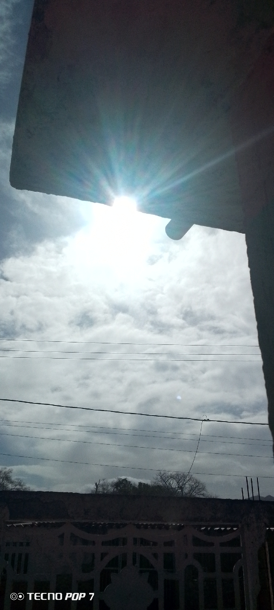 The photograph captures a bright, daytime sky with clouds, viewed from what appears to be a building in Altagracia, Venezuela. The sun is partially obscured by the building's structure, casting rays of light. Electrical power lines run horizontally across the middle ground of the frame. In the background, trees are visible over what may be a fence or wall. The bottom left corner of the image features a watermark: "Tecno Pop 7".