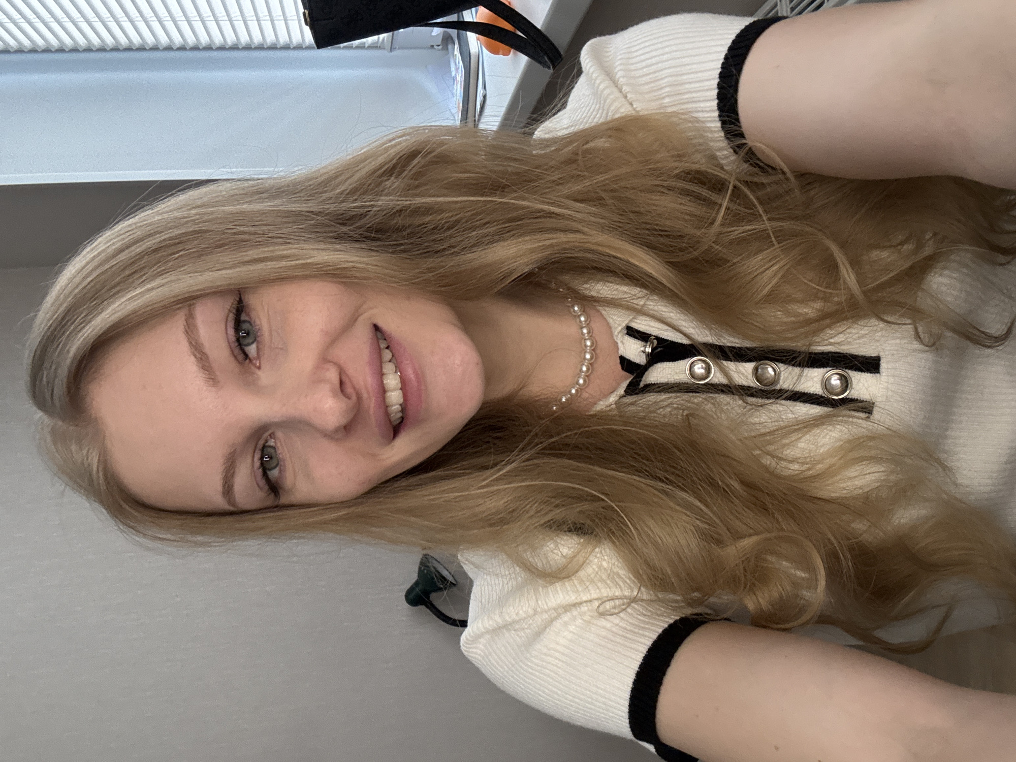 A young adult female, estimated between 20-30 years of age, is depicted in a close-up selfie-style image taken indoors in Perm, Russia. She has long, wavy, blonde hair with visible lighter roots, parted slightly off-center to her right, cascading over her shoulders. Her eyes are light-colored, appearing green or blue-grey, and she is looking directly at the camera with a wide smile, revealing her upper teeth. Her complexion is fair, with a smooth forehead and faint freckles on her cheeks. She is wearing a white ribbed short-sleeved top with black trim on the collar and sleeve cuffs, featuring a partial front placket with three round, silver-rimmed black buttons. A choker-style pearl necklace is visible around her neck, and a silver ring is present on her left ring finger. The background on her left consists of a plain light grey wall, with a small, dark green adjustable desk lamp mounted on it, positioned behind her left shoulder. To her right, a window with white horizontal venetian blinds is visible, along with a light blue wall section. On a surface below the window, several notebooks or papers and a partial view of an orange object and a black bag are discernible. The lighting appears to be a combination of natural light from the window and ambient indoor illumination.
