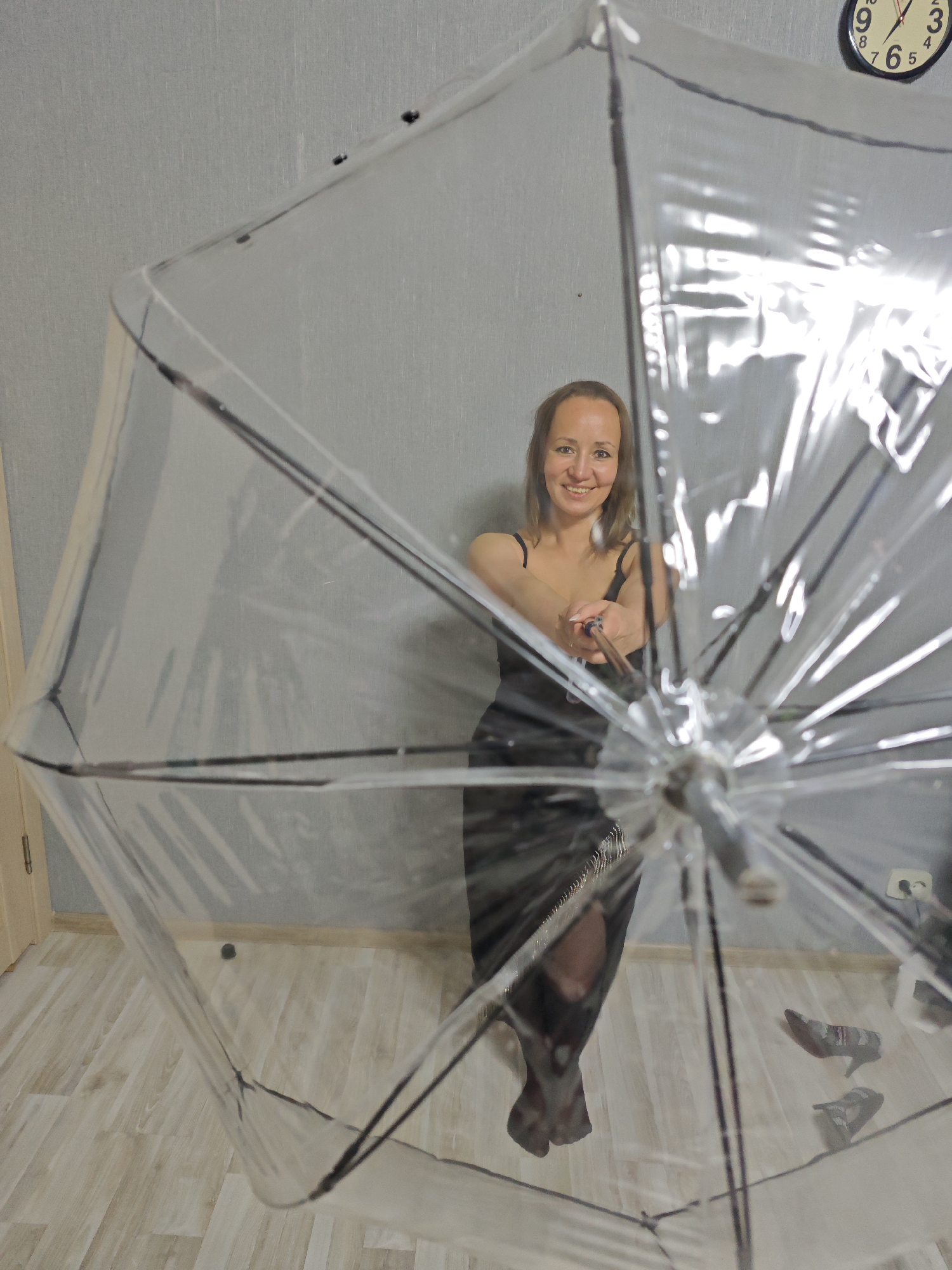 A woman stands in a room within Марковский, Russia, holding a clear umbrella. She is smiling and wearing a black dress. The room has grey walls and a light-colored wood floor. A clock is visible on the wall, and a pair of shoes and an electrical outlet are on the floor in the background.
