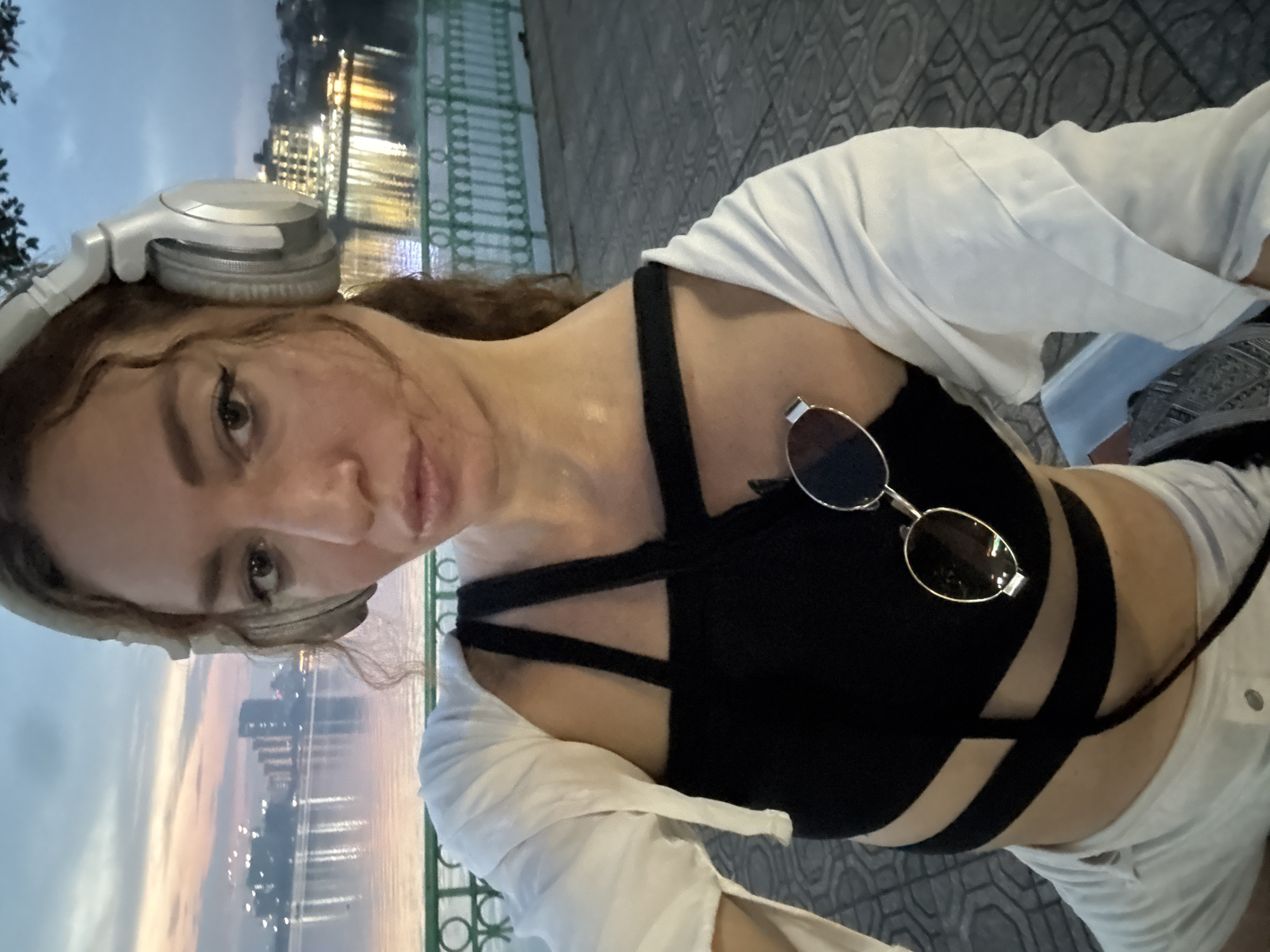 The image shows a young woman in what appears to be an outdoor setting at dusk. She is wearing a black, strappy tank top, white shorts, and an unbuttoned white shirt draped over her shoulders. Silver headphones are resting on her head, and a pair of sunglasses are clipped to her tank top. She is looking towards the camera with a neutral expression, and her makeup is visible.
In the background, there is a body of water reflecting the lights of buildings on the opposite bank. The sky shows the soft colors of sunset or twilight. A green railing and a patterned stone walkway are also visible. The overall impression is that of a casual evening scene, possibly a walk along a waterfront. The location is identified as Hà Nội, Vietnam, which aligns with the architectural style of the distant buildings and the presence of a body of water. No visible text is present in the image.