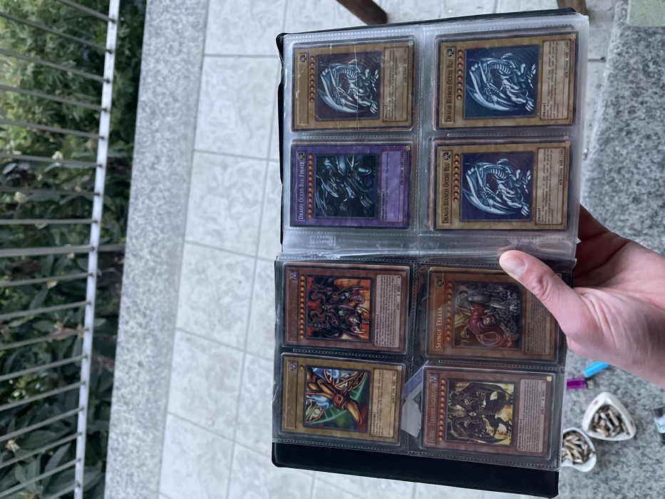 An outdoor, eye-level shot in Castione, Italy, captures a person's hand holding open a black Yu-Gi-Oh! card binder. The binder is filled with clear plastic sleeves, each holding a collectible card. The bright daylight illuminates the scene, which appears to be on a tiled balcony or patio, with a metal railing and lush greenery visible in the blurred background to the left.
The binder predominantly features Italian Yu-Gi-Oh! cards, with several copies of "Drago Bianco Occhi Blu" (Blue-Eyes White Dragon) and one "Drago Occhi Blu Finale" (Blue-Eyes Ultimate Dragon) clearly visible in the top and middle rows. Other cards include "Sfinge Teleia" (Sphinx Teleia) and a card with the visible text "SOBRE TEAMORE INCARNATA," likely a misread of "Serpent Night Dragon" or similar. The cards show some signs of use, suggesting they are part of a cherished collection.
To the right of the binder, a blue and a yellow/orange lighter are partially visible on the tiled floor, along with two small, white, shell-shaped dishes containing what appear to be cigarette butts and ash, suggesting a casual, relaxed outdoor setting. The activity depicts the act of reviewing or showcasing the card collection.