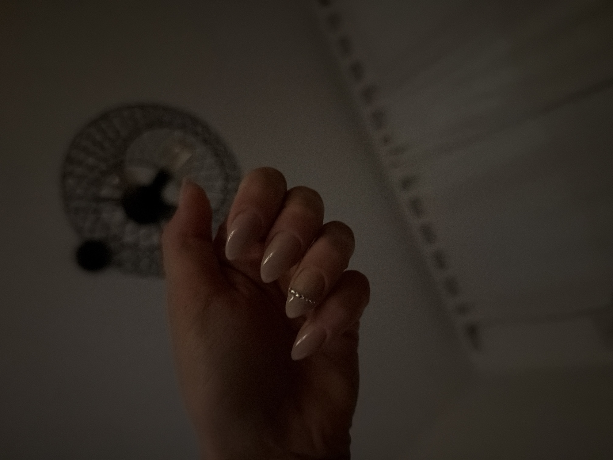 A hand with painted fingernails and a ring is in the foreground. In the background is a ceiling fan and a heating vent. The location is unknown and not specified.