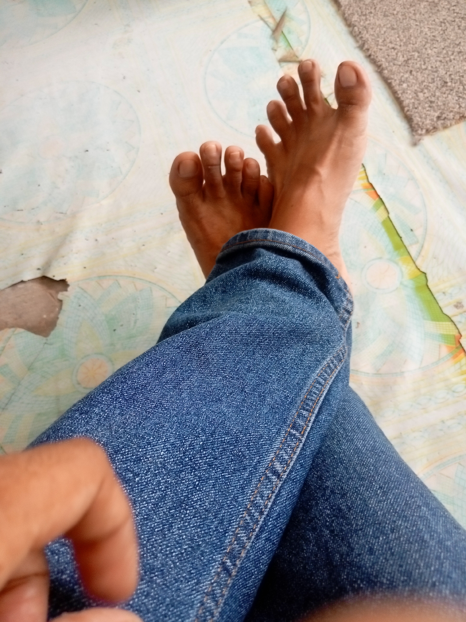 The image shows a close-up view of a person's bare feet and legs, wearing blue denim jeans. The feet are positioned on a patterned surface that appears to be a mat or floor covering with circular designs. The person's toes are spread apart. A blurry hand is visible in the lower left corner of the image. The environment is indoors, on a patterned floor. No specific location in Batam, Indonesia can be identified from the image.