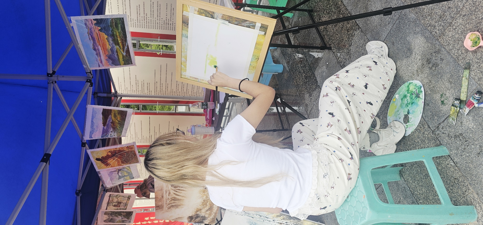 A high-angle, full shot depicts a blonde-haired woman seated outdoors, painting on an easel. She wears a plain white t-shirt, patterned light-colored pants, and white sneakers. She sits on a turquoise plastic stool with a paint palette on the ground near her feet. She holds a paintbrush in her right hand, directed towards a canvas framed on the easel.
The painting setup is under a blue canopy tent. Several landscape paintings are displayed on the left side of the canopy. To the right, behind the easel, are two vertical signs with printed text in Chinese. A clear water bottle and a smaller pink bottle are visible on a table behind the easel.
Additional painting supplies, including paint tubes, are located on the ground near the paint palette. The location appears to be a public outdoor space, evident from the gray tiled pavement visible beneath the artist's feet and the various art-related items. The scene is possibly in the city of Chongqing High-tech Zone, China, judging from the presence of Chinese text on the signs and the general environment.