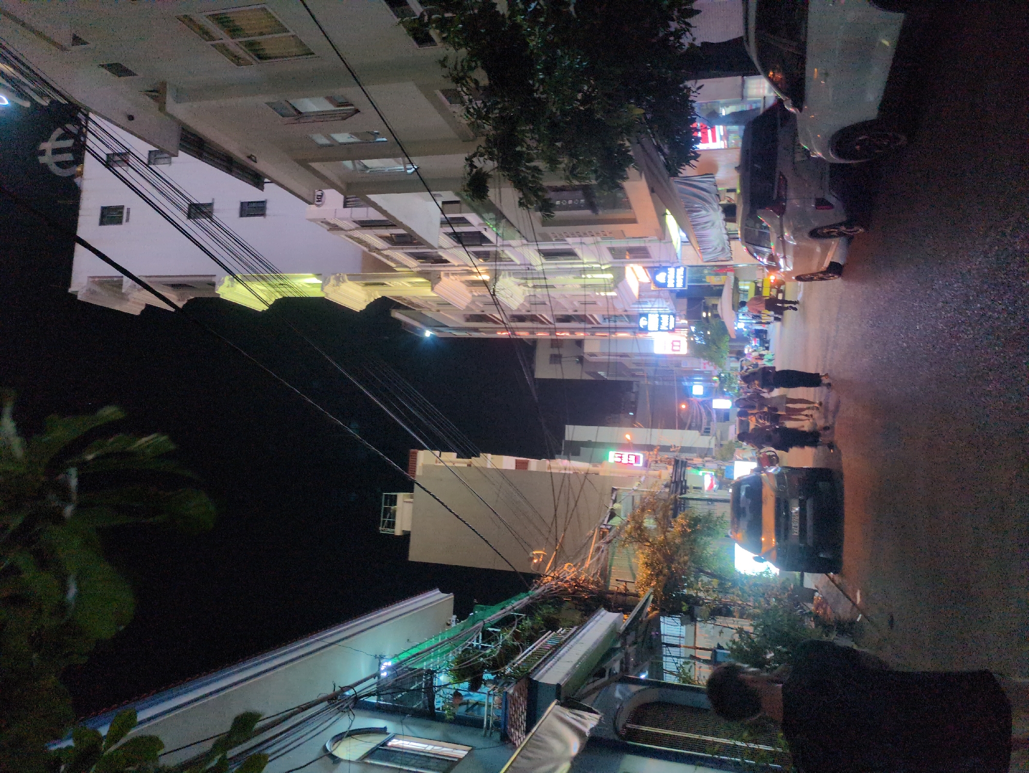 The image captures a nighttime street view within Quy Nhon Ward, Vietnam. The paved street is lined with multi-story buildings on both sides. On the left, older structures with balconies and a greenish-blue upper facade are visible, featuring extensive overhead electrical wiring. Dark tree foliage is present in the upper left foreground. On the right, taller, more modern buildings include a prominent white structure with an "HL" logo on its rooftop and several businesses displaying illuminated signs, notably "DOBAL HOTEL," along with various neon and LED lights. A dense network of black electrical wires is suspended overhead across the street.
In the immediate foreground on the left, the back of an individual wearing a dark short-sleeved shirt and light-colored lower garments is visible, facing away from the camera. In the mid-ground, a light orange/tan SUV, possibly a Chevrolet Colorado, with a partial license plate "27A-02xxx," is positioned on the left side of the street. Further down the street in the center-right, a group of approximately 5-6 individuals is walking. On the right side of the street, a white SUV, resembling a Mitsubishi Xpander, is parked, facing away. The front of another white vehicle is partially visible parked further ahead. Another individual in a light-colored shirt is walking on the right side of the street, and a small, light-colored utility vehicle with an umbrella-like cover is visible in the distance. The street is illuminated by ambient light from building facades, signs, and streetlights, creating reflective patches on the road surface. Trees are present along the sidewalks.