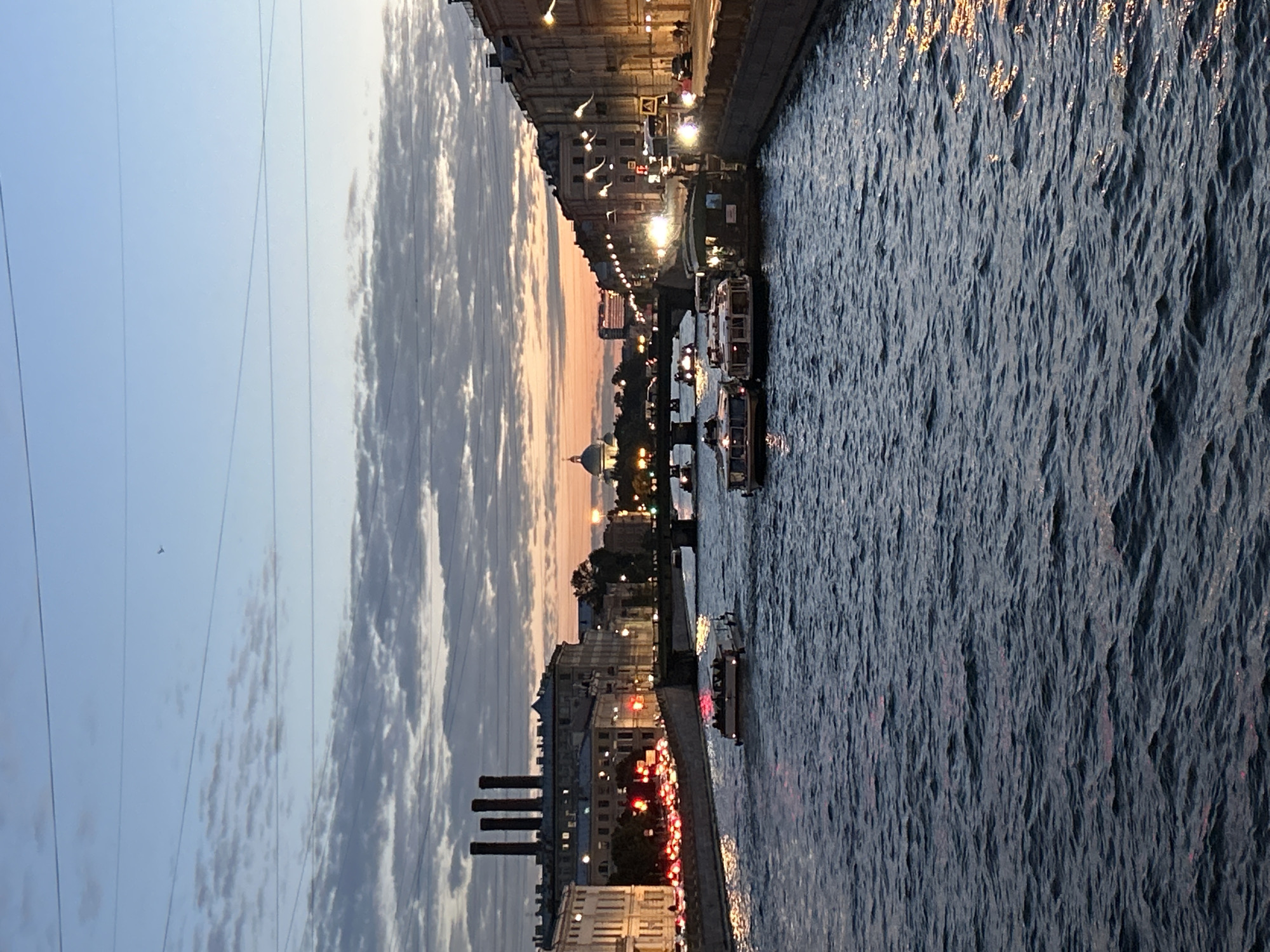 The image depicts a canal in a city at dusk. Several tour boats are navigating the waterway, filled with passengers. The canal is lined with buildings, some featuring illuminated windows and streetlights. In the distance, a large dome is visible, suggesting a prominent landmark such as a cathedral or government building. The sky is a gradient of blue and orange, with scattered clouds, indicating the end of daylight. Overhead, power lines crisscross the sky, and a single bird is visible in flight. The water surface shows reflections of lights from the buildings and boats. The overall scene conveys a sense of urban tranquility as evening sets in.