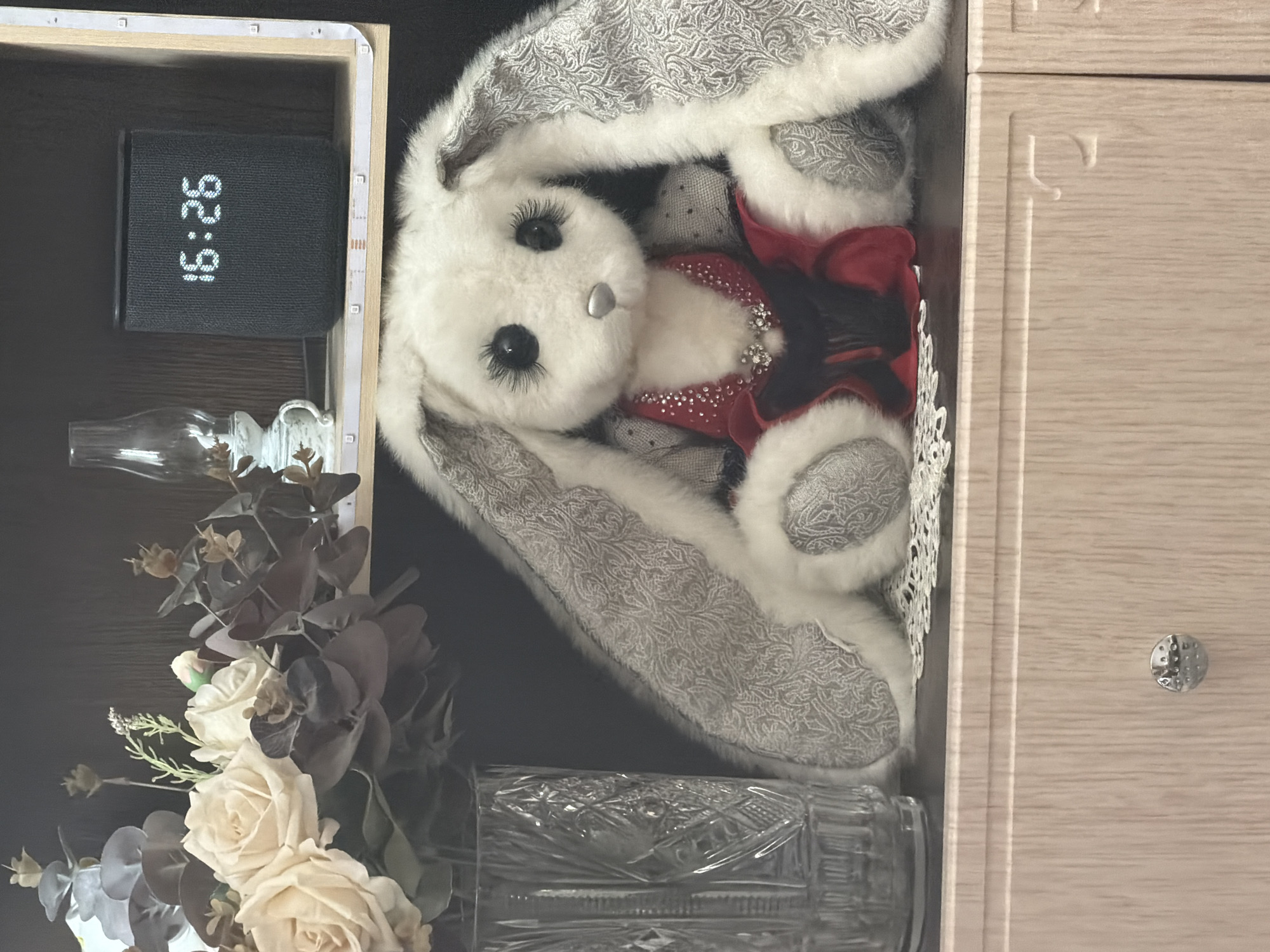 A stuffed white bunny with long ears and large, dark eyelashes sits in the center of the image, dressed in a red and black outfit adorned with rhinestones. The bunny is positioned on a shelf or tabletop, in front of a floral arrangement of cream-colored roses and dark leaves in a clear, ornate glass vase. To the right of the bunny, a black speaker with a digital clock displaying "16:26" is visible. Above the speaker, a small glass lamp with a white base sits on a shelf. The background appears to be a dark wooden shelf. Below the bunny is a light-colored wooden drawer with a silver knob. The scene is indoors, likely a domestic setting. There is no discernible connection to Nizhnevartovsk, Russia.