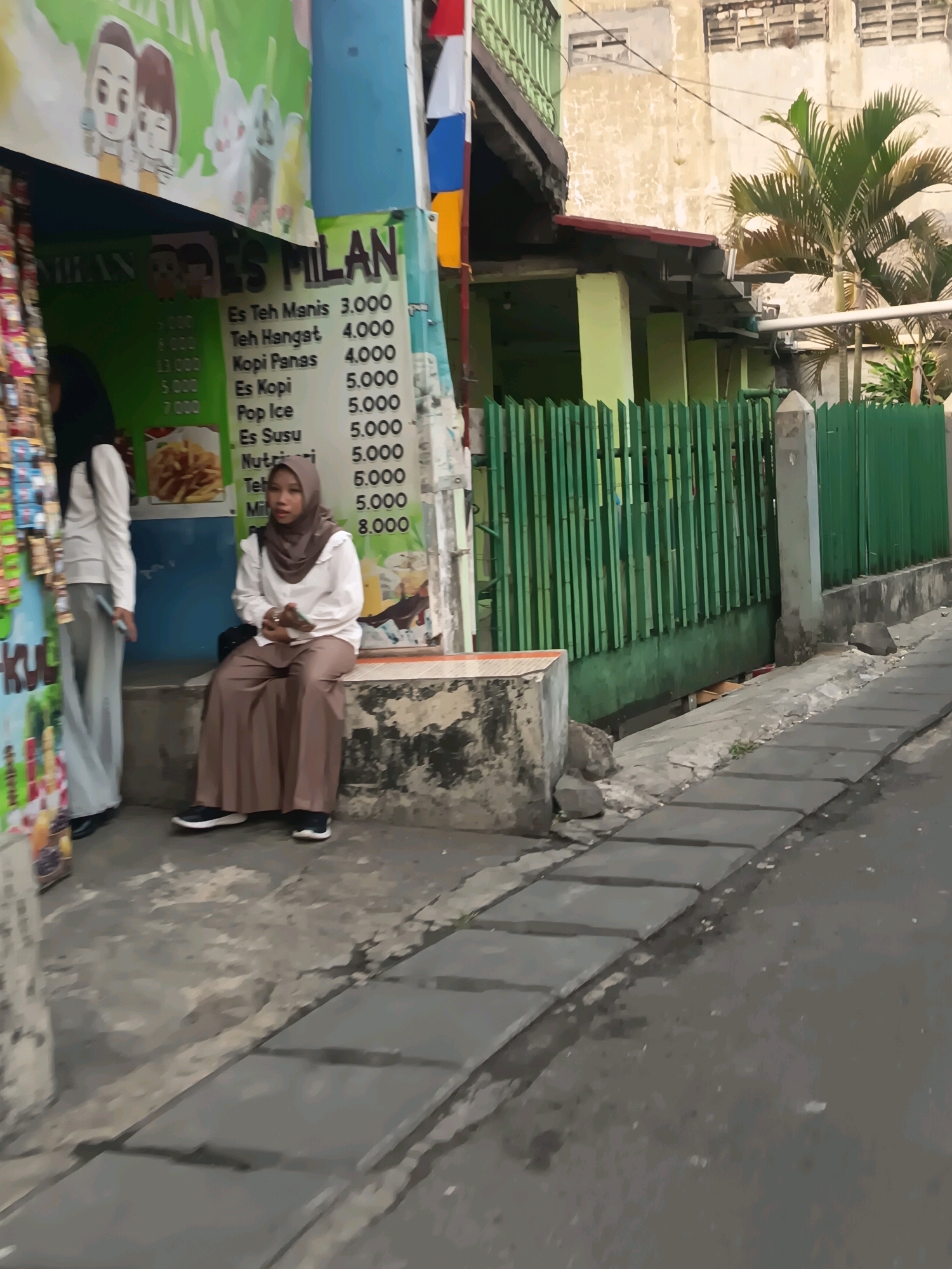 The image captures a street scene in Tangerang, Indonesia, focusing on a small food stall. In the foreground, a young woman wearing a hijab and a white shirt with a ruffled collar, paired with brown pants and black shoes, is seated on a concrete bench. She holds a small object in her hands. To her left, another woman in a white shirt and grey pants, also wearing a hijab, stands near a display of products.
Behind them, a stall displays a menu board with items like "Es Teh Manis," "Teh Hangat," "Kopi Panas," "Es Kopi," "Pop Ice," "Es Susu," and "Nutrijell," along with prices. Above the menu, a colorful banner features cartoon characters. The stall is situated alongside a residential building with green wooden fencing and a palm tree visible in the background. The pavement in front of the stall is made of grey square tiles, and the street is paved with asphalt. The overall impression is of a bustling urban environment with everyday commerce.