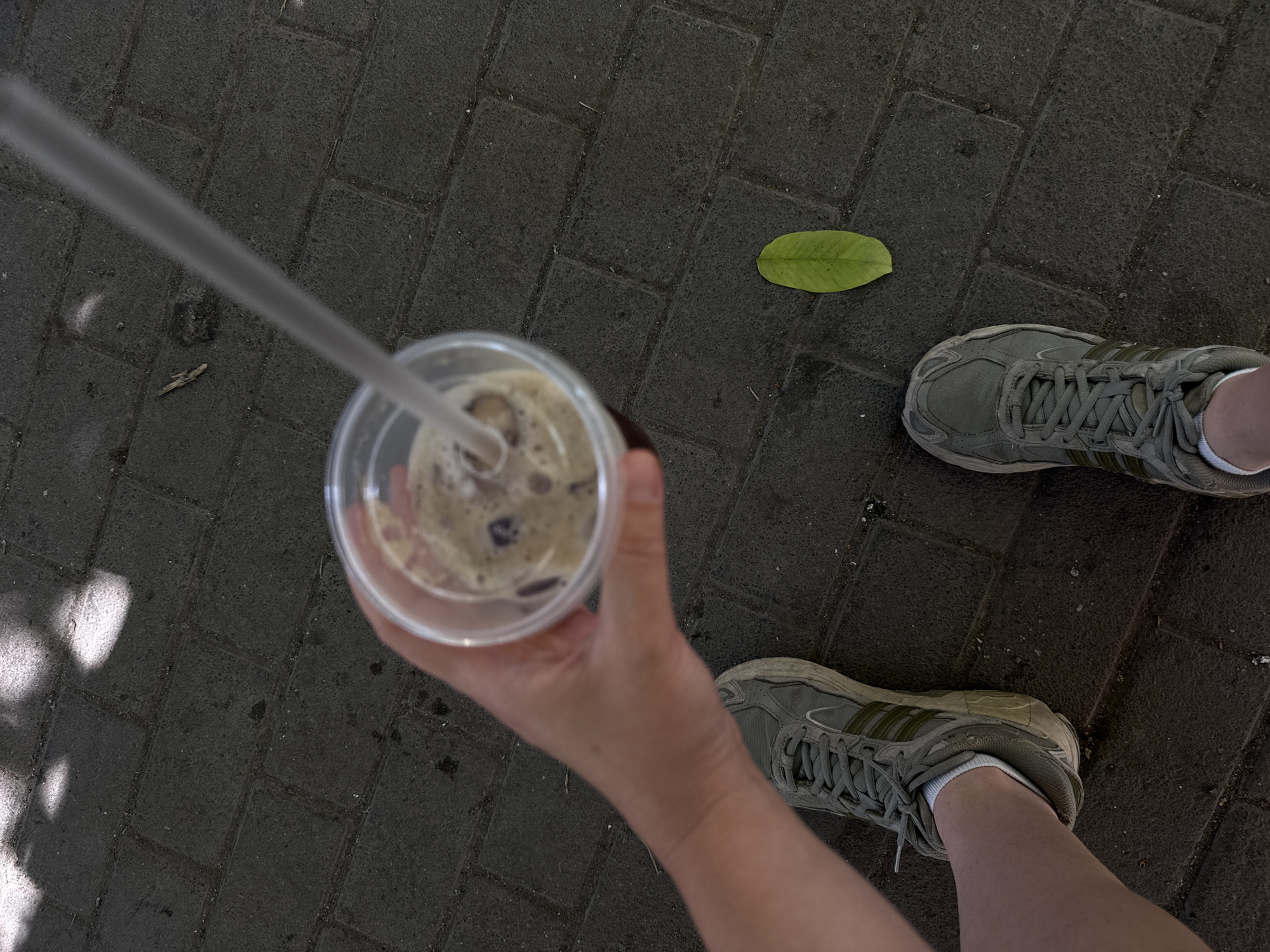 An over-the-shoulder point-of-view shot captures a person's hand holding a clear plastic cup filled with an iced coffee drink. The cup, complete with a clear plastic straw, is held in the foreground. The person is wearing grey sneakers with white socks. A single green leaf rests on the dark grey brick pavement. Sunlight casts dappled shadows from unseen trees onto the pavement, suggesting an outdoor urban setting. The scene is consistent with an outdoor cafe or public walkway in Yerevan, Armenia, possibly in a park or pedestrian area.