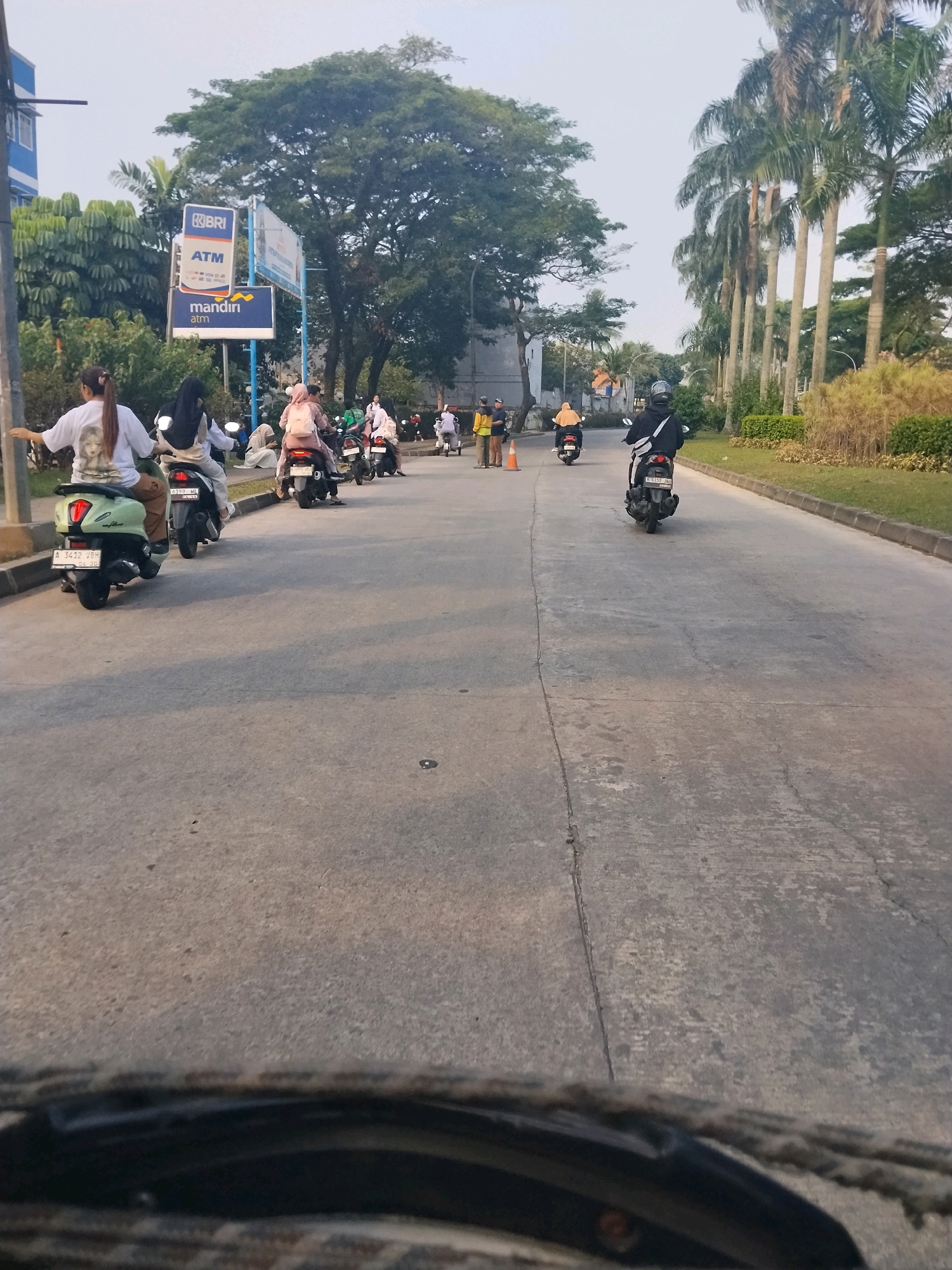 The image shows a street scene in Mekar Bakti, Indonesia, with several people riding motorcycles. In the foreground, a light green scooter with a license plate reading "A 3412 VBH" is visible from the rear. The rider is wearing a white shirt with a graphic print of a person's face. Behind this scooter, another scooter, a dark-colored one with a rider wearing a black hijab and backpack, is also seen. Further down the road, a pink-clad rider on a scooter is approaching. To the left of the road, a sign with the BRI logo and "ATM" is displayed, along with a "mandiri atm" sign below it. Tall palm trees line the right side of the road, and lush green vegetation is present. The road itself is paved with concrete and shows some wear and tear. The sky is overcast. The scene appears to be a public road in an urban or semi-urban area.
