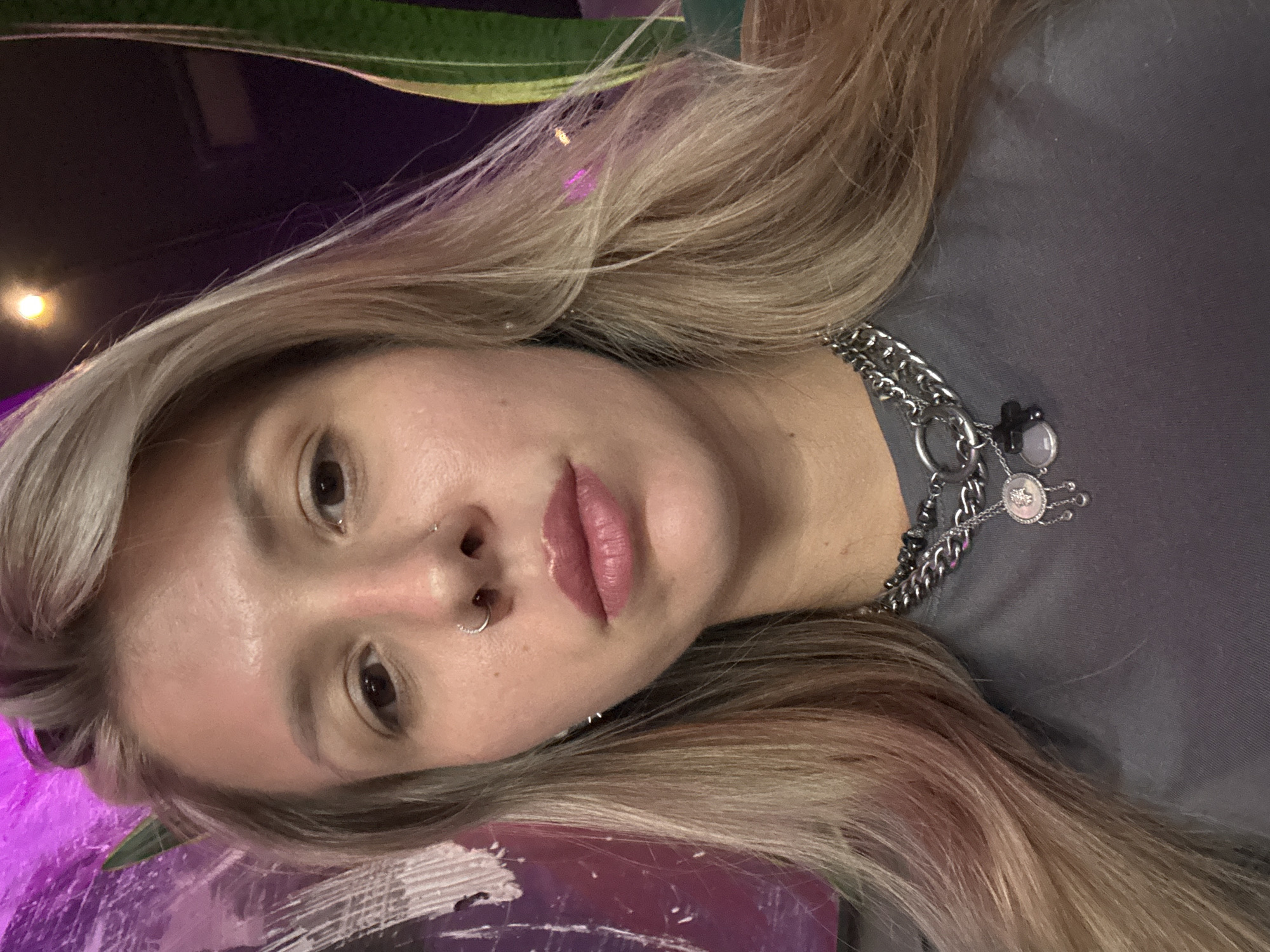 A medium shot, waist-up, selfie-style photograph shows a woman with blonde hair and light skin, looking directly at the camera. She is wearing a gray t-shirt and a multi-layered silver chain necklace with a circular pendant featuring dangling elements and a small black cross. She has a nose ring and appears to be indoors in an area with purple and dark backgrounds, with a hint of green foliage on the right side of the frame. The lighting is dim with some purple and yellow hues reflecting on her hair.
