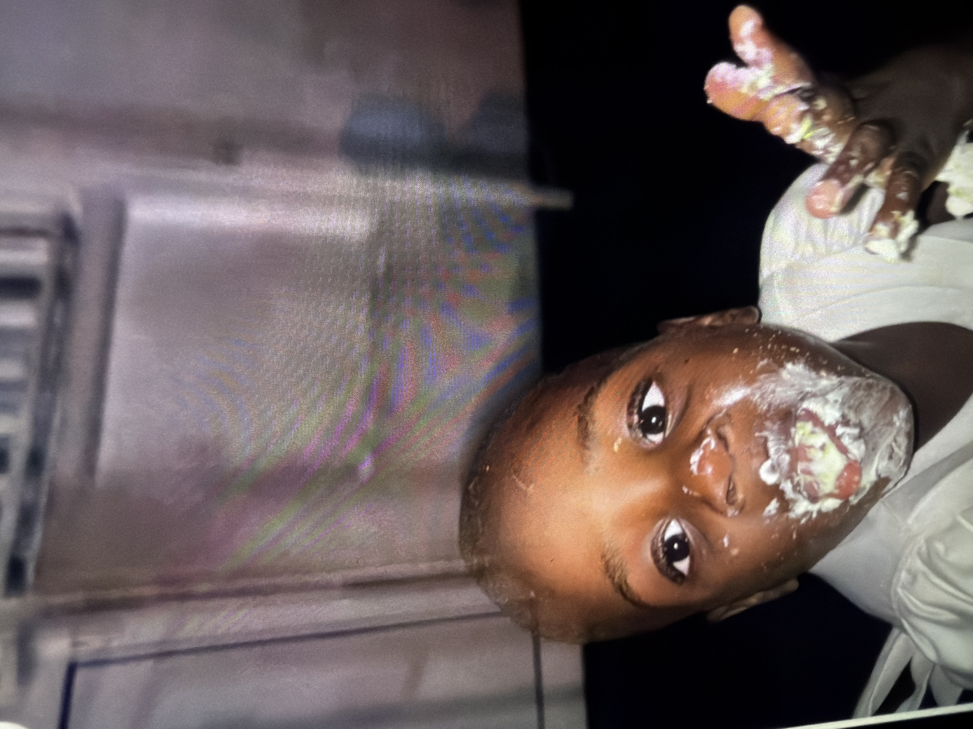 A young child with dark skin and short, dark hair is shown close-up, looking towards the camera with wide eyes. The child's face and hands are smeared with white frosting and possibly cake. The child is wearing a white shirt. The background is dark and out of focus, with some blurry, colorful streaks of light and what appears to be a wall or structure with vertical lines. The lighting is dim, and the image quality is somewhat grainy, suggesting it may have been taken at night or in low light conditions with a digital camera. The child's expression is one of innocent engagement with the frosting. The image appears to be a casual, candid snapshot.