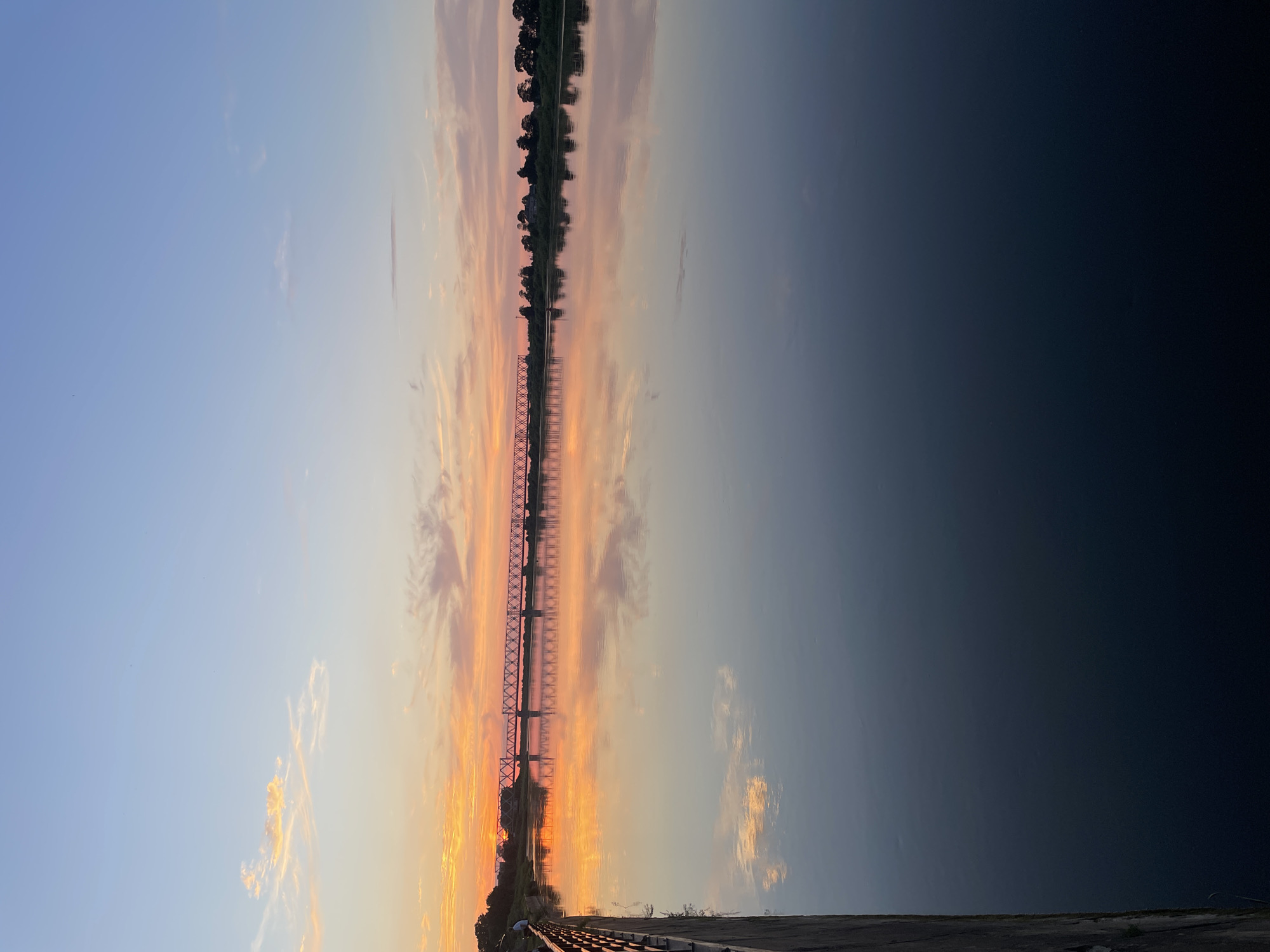 This is a vertical, eye-level, medium shot of the Sovetsk Railway Bridge over the Neman River at sunset. The sky is a gradient of soft blues, pinks, and oranges, with wispy clouds reflecting on the calm water. The bridge, a prominent steel structure with intricate lattice work, stretches across the horizon. The far bank of the river is lined with trees and some distant buildings, all silhouetted against the colorful sky and mirrored in the water. On the left side of the frame, a dark, textured wall or embankment with a railing runs along the edge of the river. A single person is visible on the embankment, looking towards the sunset. The overall mood is serene and picturesque, capturing the beauty of the natural landscape and the industrial element of the bridge at dusk.