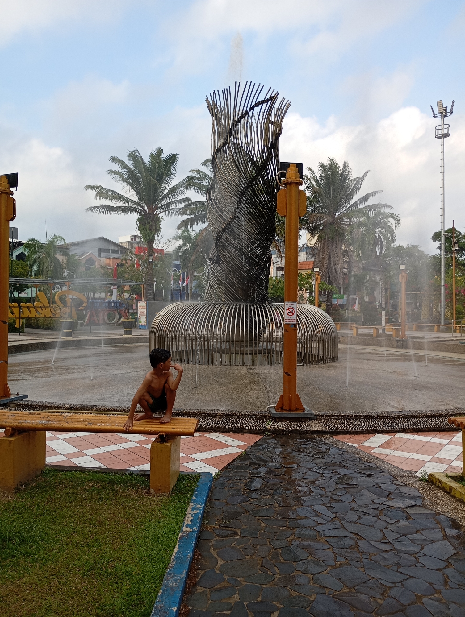 In Balikpapan, Indonesia, a young boy is crouched on a bamboo bench in a public plaza. The plaza features a large, sculptural fountain with a central, twisted metal structure and a circular base with metal bars. Water sprays from several nozzles around the fountain, creating a misty atmosphere. The boy is shirtless, wearing dark shorts, and appears to be looking to his right. Behind the fountain, palm trees and buildings are visible. The ground is a mix of wet paving stones and a patterned tiled area. A tall light pole stands to the right of the fountain. The sky is partly cloudy. A "No Smoking" sign is affixed to one of the fountain's support poles.