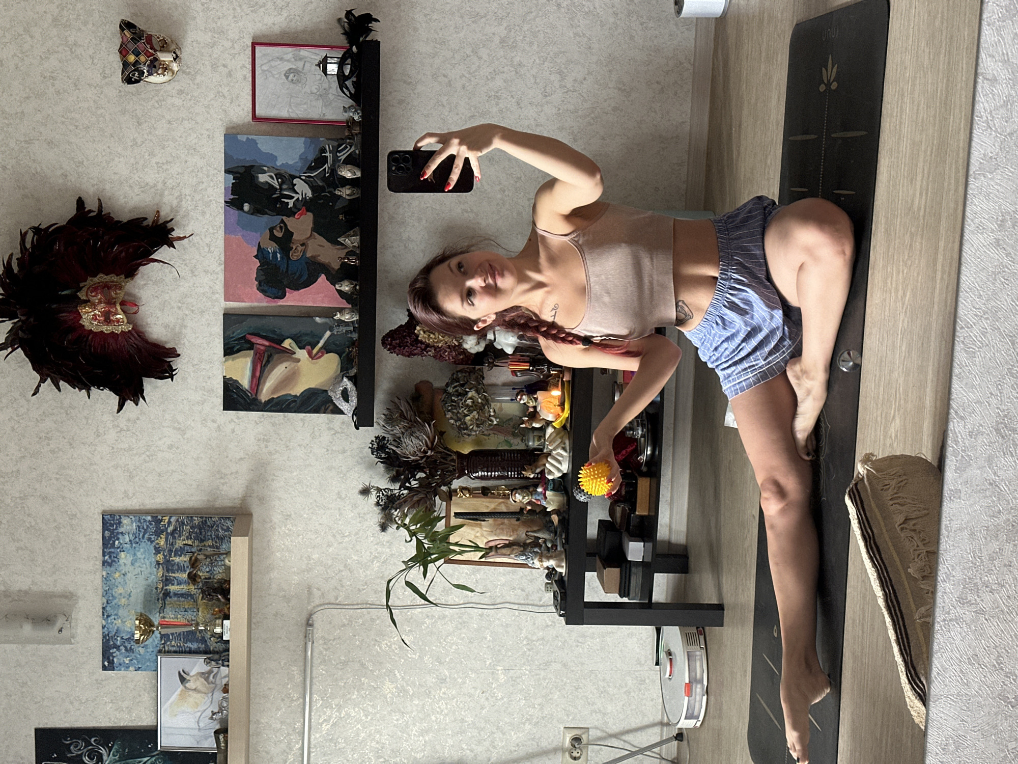 A woman with auburn hair braided to her left side and a tattoo on her left hip is performing yoga on a black yoga mat in what appears to be her living room. She is wearing a tan tank top and blue striped shorts. She is holding a yellow massage ball in her right hand, and her phone in her left hand, taking a selfie. On the floor to her left is a robotic vacuum cleaner. The room is decorated with various items, including paintings on the wall, figurines on shelves, and dried floral arrangements. One painting depicts a woman smoking a cigarette, another shows Batman and Catwoman, and a third is a sketch of a woman. A red feathered headdress and a mask are hung on the wall above the paintings. The room has textured grey wallpaper and light wood-paneled floors. The scene is lit by overhead lighting and natural light coming from a window that is not visible in the frame. The general decor and style of the apartment suggest it is located in Russia, though no specific indication of the city of Vladimir is present within the image itself.