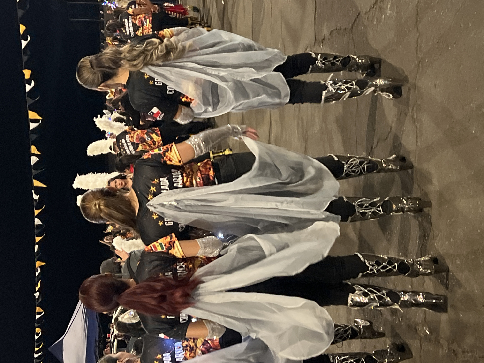 Arica, Chile: A sideways view of a procession of participants, most likely from a school, wearing matching black tops and pants. Some are adorned with a white feather headpiece. All have silver mid-calf boots with ornamental criss-cross embellishments. All are wearing sheer capes that appear to be backlit. Bunting is visible on the left side. The environment appears to be an outdoor street at night, illuminated with artificial light.