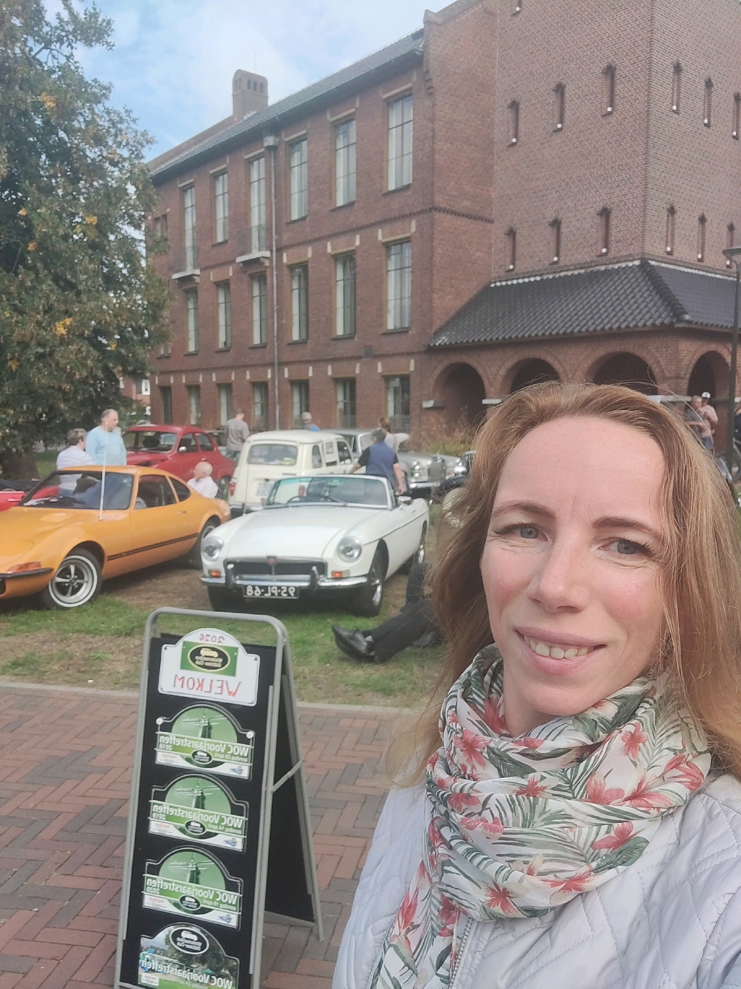 A woman with blonde hair and a floral scarf smiles at the camera in the foreground, with a classic car show in the background. The event appears to be taking place in Winterswijk, Netherlands, suggested by the Dutch text on the signage. The background features several vintage cars, including a bright orange sports car and a white convertible. A brick building, possibly a historical site or public building, serves as the backdrop. The scene is set outdoors on a paved area with grass. People are visible milling around the cars, suggesting a public gathering or exhibition. The lighting indicates it is daytime, with overcast skies.