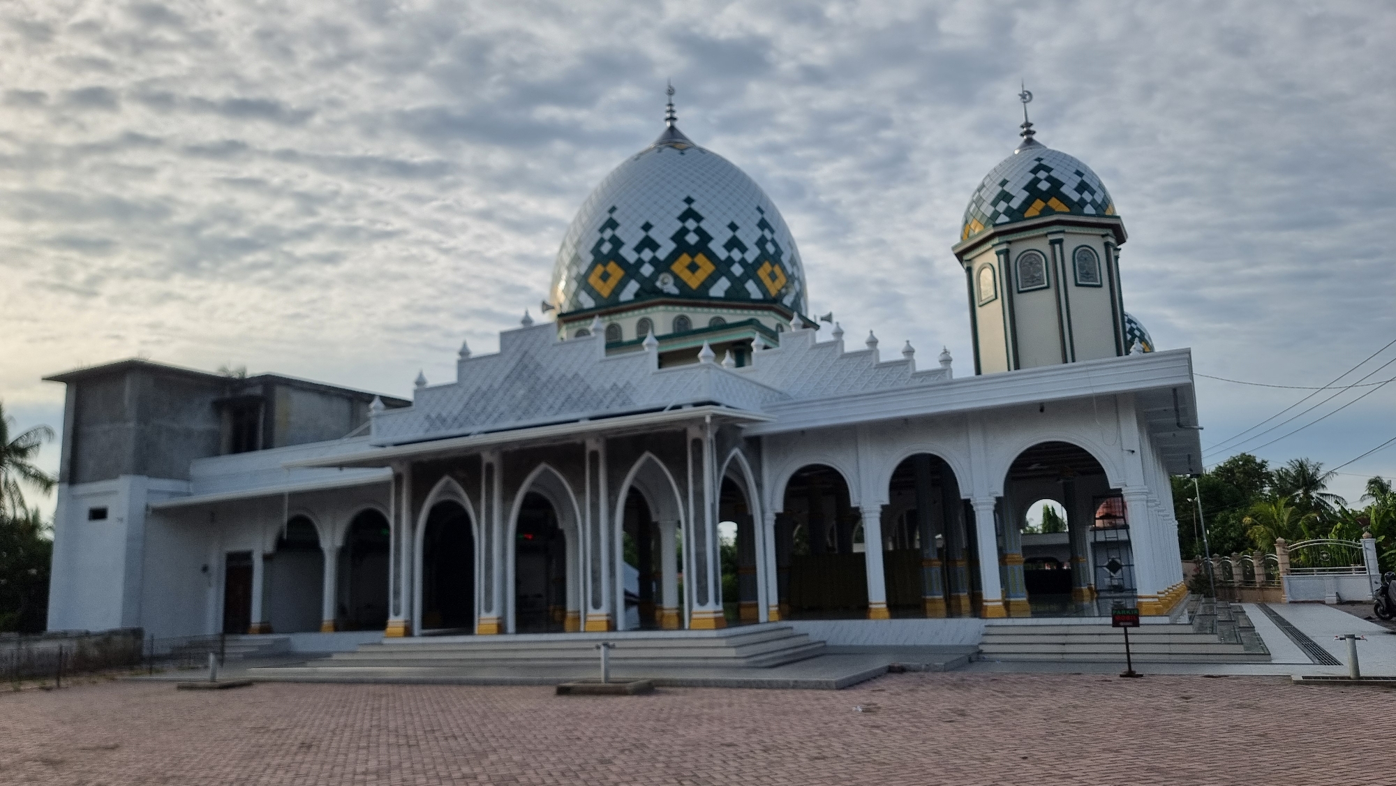 The image shows a mosque in Meunasahlhok, Indonesia. The mosque features a large central dome and a smaller dome on a minaret, both adorned with a geometric pattern of green, yellow, and white tiles. The main building is white with arched walkways and decorative pillars. A paved courtyard with red brick pavers is in front of the mosque. The sky is overcast with scattered clouds. The surrounding area shows some vegetation, including palm trees, and a fence in the background. There are no people visible in the image.
