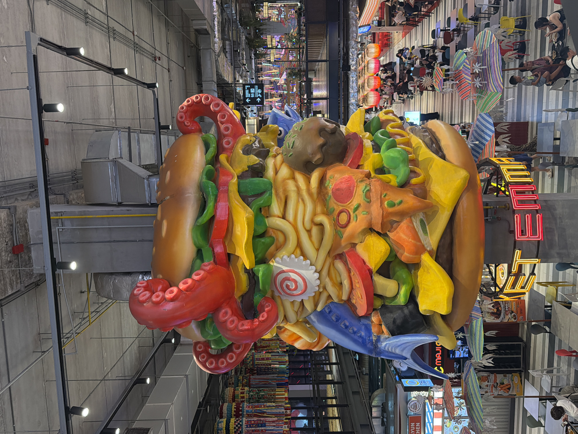The image captures a vibrant indoor scene, likely a shopping mall or food court, dominated by a large, colorful, and highly detailed food sculpture hanging from the ceiling. The sculpture is shaped like a giant burger or sandwich bun overflowing with an eclectic mix of exaggerated food items.
In the foreground, the massive sculpture features prominent red octopus tentacles emerging from the bun, alongside thick yellow noodles, green peppers or vegetables, red tomato or salami slices, yellow chunks resembling cheese or pineapple, a mushroom-like object, and distinct pizza slices with pepperoni. A blue, fish-like element is also visible, along with a white disc patterned with a red swirl, reminiscent of narutomaki. The sculpture's intricate details and bright colors make it a striking focal point.
In the background, multiple levels of the building are visible. A dining area on an upper level shows numerous people seated at tables and chairs, some of which are brightly colored. Striped sun umbrellas add to the casual, lively atmosphere. Decorative orange and red lanterns hang above some of the dining spaces. Further back, more people are visible, some standing, some seated.
Architectural details include an industrial-style ceiling with exposed ventilation ducts, pipes, and track lighting fixtures. A black directional sign with white arrows points to facilities like toilets, escalators, and a baby changing station. Several neon signs are scattered throughout the background, including one that appears to spell out "FOOD" in red and orange, and another smaller sign with partial text that reads "N.KITCHEN". The overall setting suggests a bustling commercial area, possibly a food-themed zone within a larger complex, designed to be visually engaging and attractive to visitors. The lighting indicates an indoor setting, likely during daytime or well-lit evening hours.