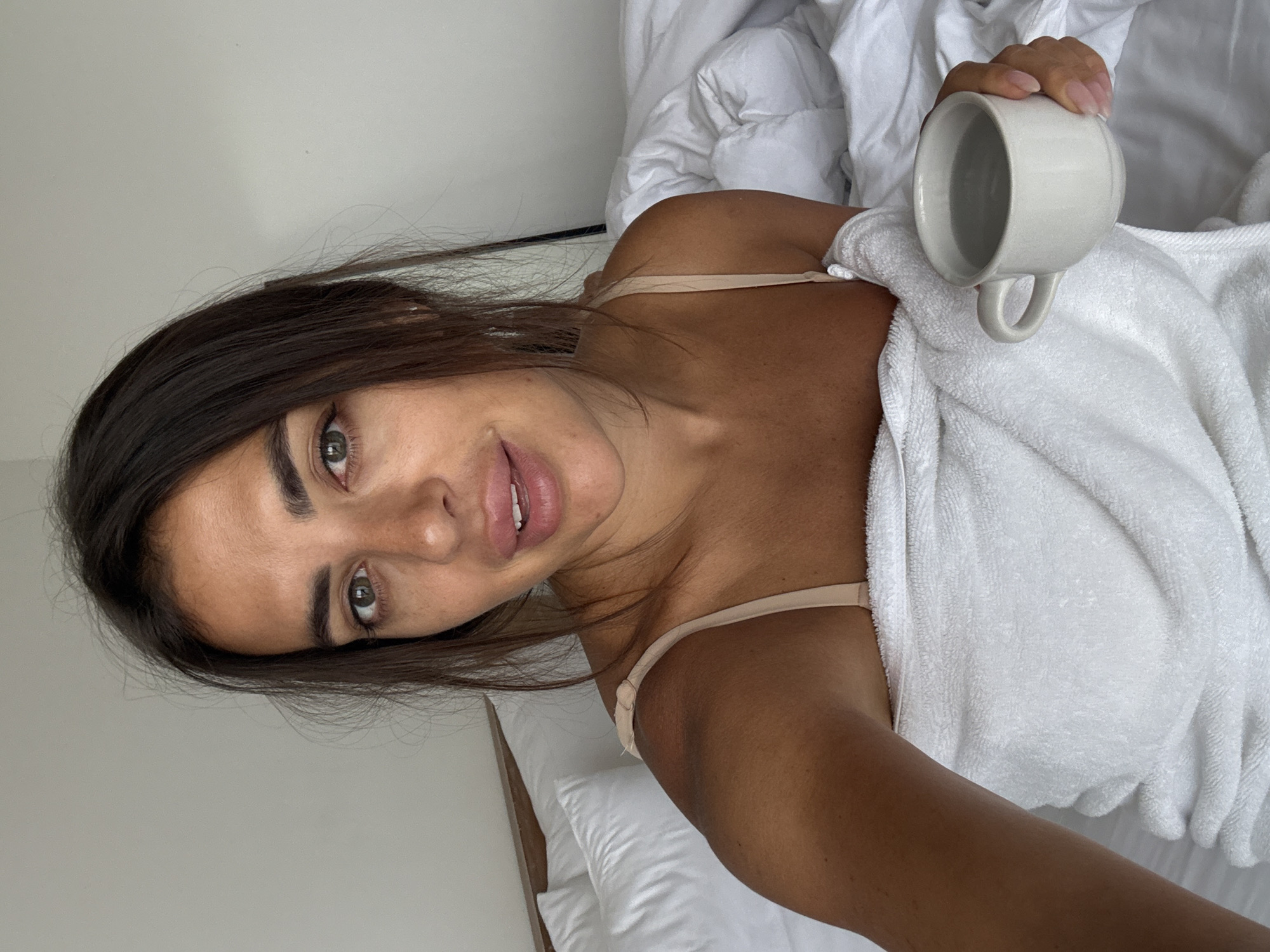 The image displays an adult woman in a bedroom setting in Kuta Selatan, Indonesia. She is depicted from the chest up, capturing a selfie-style photograph. Her dark brown hair falls loosely around her face, and her eyes, appearing greenish-hazel, are directed straight at the camera. She has a tanned complexion with visible freckles across her nose and cheeks, and her lips are full with a subtle, relaxed expression. She wears a thin-strapped, nude-colored bra, with her upper body wrapped in a white terry cloth towel. In her right hand, she holds a plain white ceramic mug filled with a translucent, light-colored liquid, likely water. She is seated in a bed featuring clean white sheets and a fluffy white duvet. A simple, light-colored wooden headboard is visible behind her left shoulder, and the background consists of a plain, light-toned wall. The lighting is soft and even, suggesting an indoor, likely morning, environment.