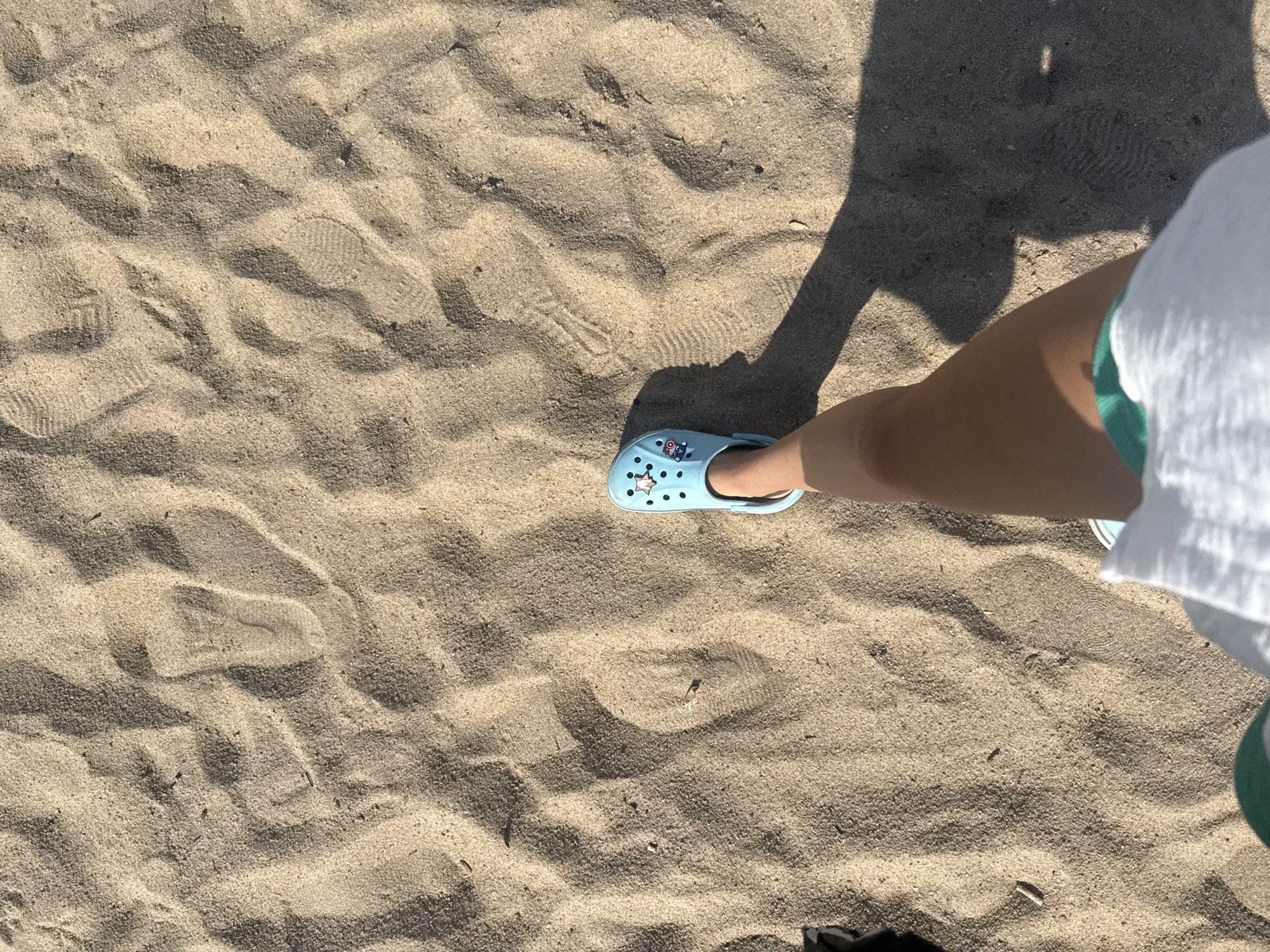 The image captures a first-person perspective of someone walking on a sandy surface, likely a beach. The sand is light brown and textured, with visible ripples and footprints. A single light blue Croc shoe with cartoon charms is worn on the person's left foot, which is clearly visible. The person's leg, from the knee down, is also in view, wearing the shoe and what appears to be white shorts with a green waistband. A long shadow stretches from the person's leg and shoe across the sand, indicating a sunny day. The scene is devoid of any specific landmarks or clues that would definitively place it in Nha Trang, Vietnam. However, the overall environment of sand and beach is consistent with coastal areas.