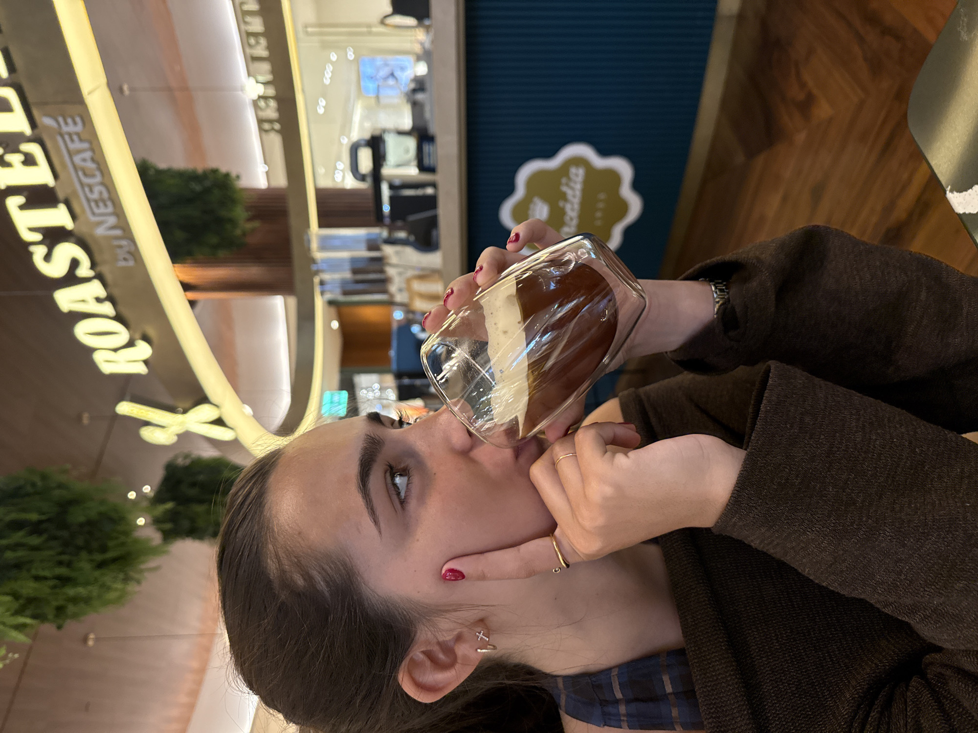 A close-up, horizontally oriented shot depicts a young woman in an indoor café, seen from a side profile. She is actively drinking from a clear, square-shaped glass filled with a layered beverage, appearing to be an iced coffee with a creamy or foamy top. Her gaze is directed upwards and slightly to the left, beyond the frame.
The woman is wearing a dark brown, possibly knit or wool, jacket over a plaid shirt. Her right hand, with red nail polish and a gold ring, is positioned near her chin. A silver-toned wristwatch is visible on her left wrist. She has dark hair, styled away from her face, and small earrings, one appearing to be a cross and the other a hoop.
The background is a modern indoor setting, likely a café or a commercial space. A prominent, backlit sign with gold lettering clearly displays "ROASTED by NESCAFE," indicating the establishment's affiliation. Further to the right, a dark teal wall features an ornate gold logo with visible text, partially reading "acídia," which, in the context of Matosinhos, Portugal, strongly suggests the brand "Arcádia," a Portuguese chocolate and coffee house. Green decorative plants or small trees are also visible in the softly blurred background. The overall lighting is bright and even, consistent with an indoor daytime or well-lit evening environment.