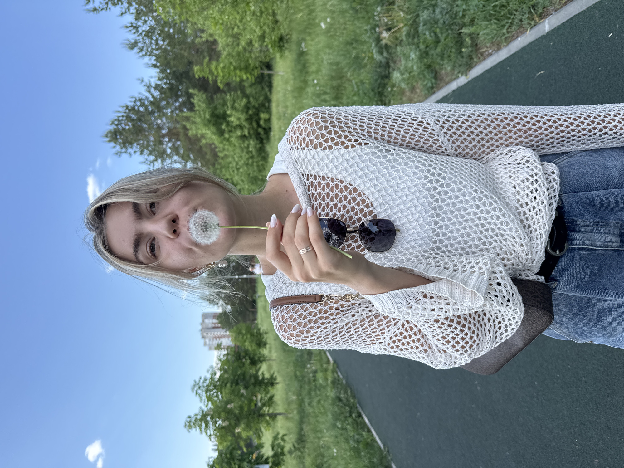 A young woman with blonde hair is seen outdoors, holding a dandelion to her lips as if she is about to blow the seeds. She wears a white knitted sweater over a white top, and blue jeans with a dark belt. Sunglasses are clipped to her sweater. She has a ring on her finger. In the background, there is a green, grassy area with trees and, in the distance, a multi-story building can be observed. The area appears to be a park or green space within an urban environment. The location is identified as Unknown, Unknown.