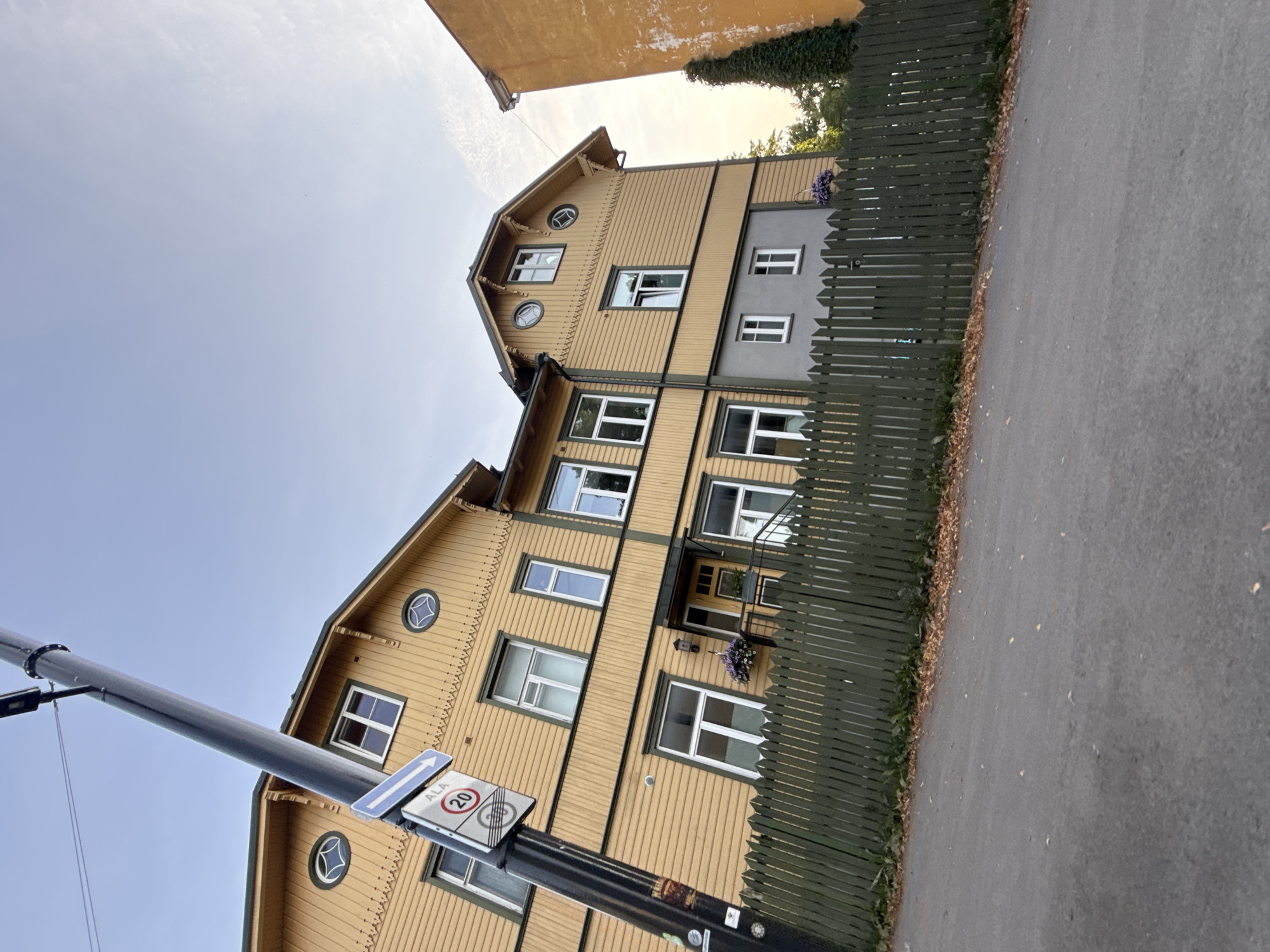 An exterior, low-angle, wide shot captures a yellow wooden building with a green fence and a street in front of it, in Tallinn, Estonia. The sky is a clear, light blue with some wispy clouds on the right side. The building has several windows with white frames and is adorned with decorative trim. A traffic sign indicating a 20 km/h speed limit is attached to a dark, vertical pole in the foreground. The fence is made of dark green wooden planks and runs along the front of the property. A grey-colored section is visible on the lower part of the building, behind the fence. The street is paved and appears to be slightly wet. Fallen leaves are scattered on the ground near the fence and on the street. A portion of an orange building is visible on the right side of the frame.