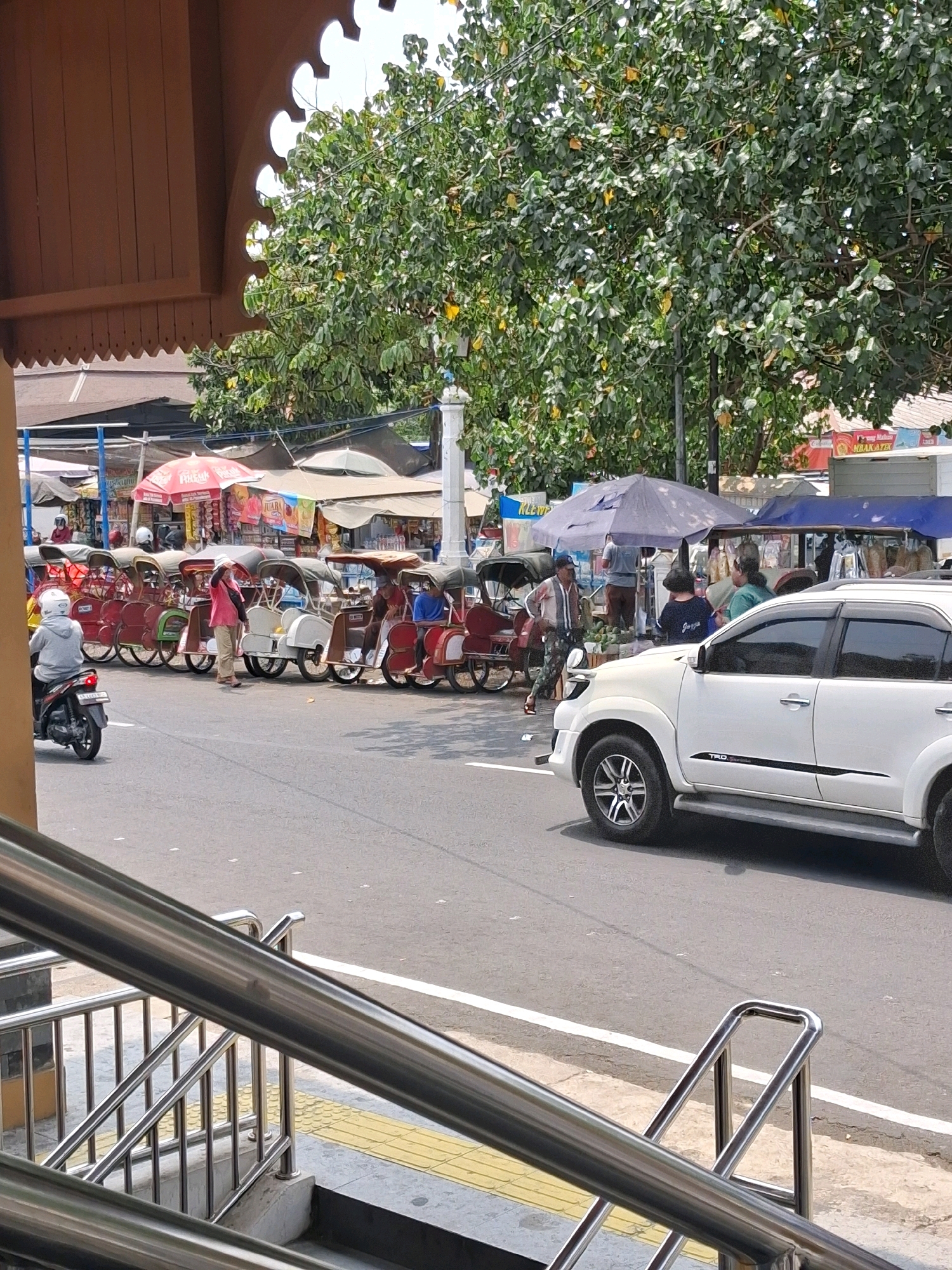 A street scene in Surakarta, Indonesia, features a line of parked becaks, or bicycle rickshaws, on the side of a paved road. A white SUV is driving on the road, passing the parked becaks. Several people are standing near the becaks, and some are sitting in them. Behind the becaks, there are market stalls with awnings and umbrellas, selling various goods. A large tree with green leaves provides shade over part of the scene. In the foreground, a metal handrail and stairs are visible. The overall impression is that of a busy street market on a sunny day.