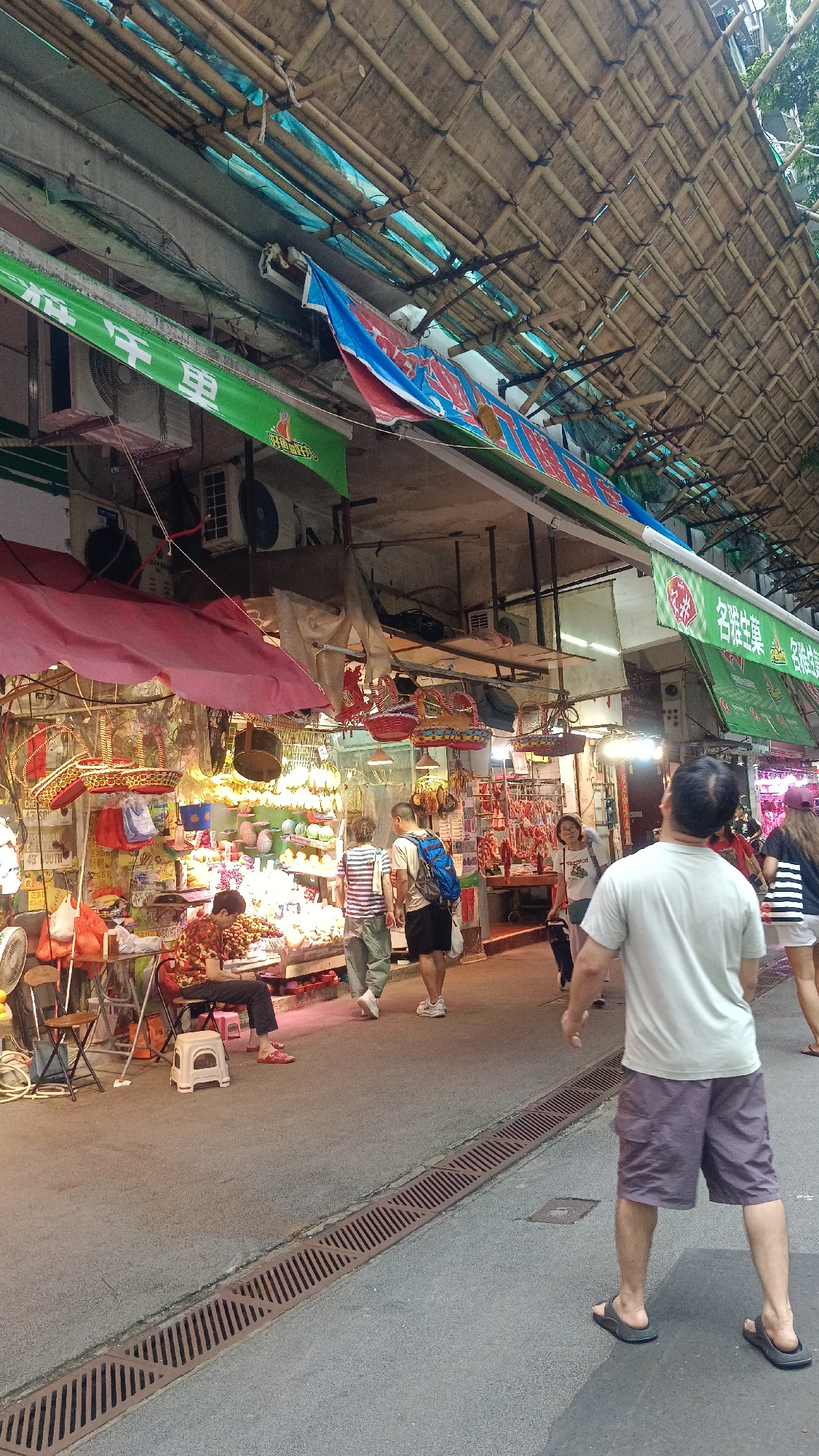 This image depicts a street market scene, likely in Hong Kong, China, under a bamboo-covered canopy. In the foreground, a person wearing a light grey t-shirt and purple shorts walks away from the camera. The street itself is paved with asphalt and features a drainage grate.
Further into the market, several stalls are visible. One stall on the left displays red awnings, with a person in a floral shirt sitting at a table filled with goods. In the middle ground, two men are walking past a stall laden with produce. The man on the left wears a striped t-shirt and light-colored pants, while the man on the right has a blue backpack and dark shorts.
Various market goods are displayed, including fruits, vegetables, and what appear to be dried meats or seafood hanging from the stalls. Colorful baskets and bags are also present. Overhead, bamboo structures and tarps create a rustic roofing system for the market. The overall impression is a bustling, traditional street market environment.