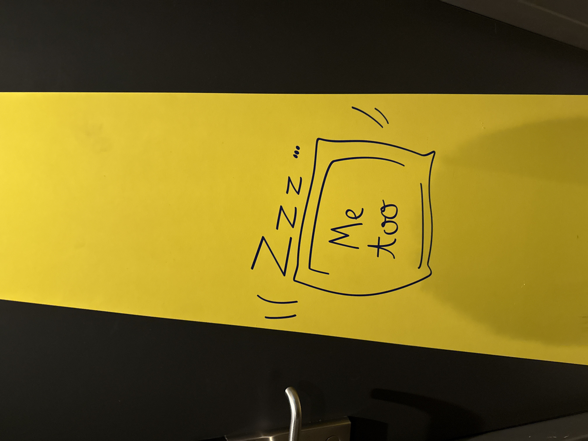 The image shows a yellow surface with a black border, likely a door or wall in a building. A handwritten message in blue ink reads "Zzz" with three dots, and a drawing of a pillow with "Me too" written inside it. There are also two curved lines on either side of the drawing, possibly representing sound waves or movement. A silver door handle is visible on the black border on the left side of the image. The background is black, suggesting the subject is partially in shadow or against a dark surface. There are no people visible in the image. The location is identified as Stein, Netherlands, although the specific context of this message and drawing within the city is not provided by the image.