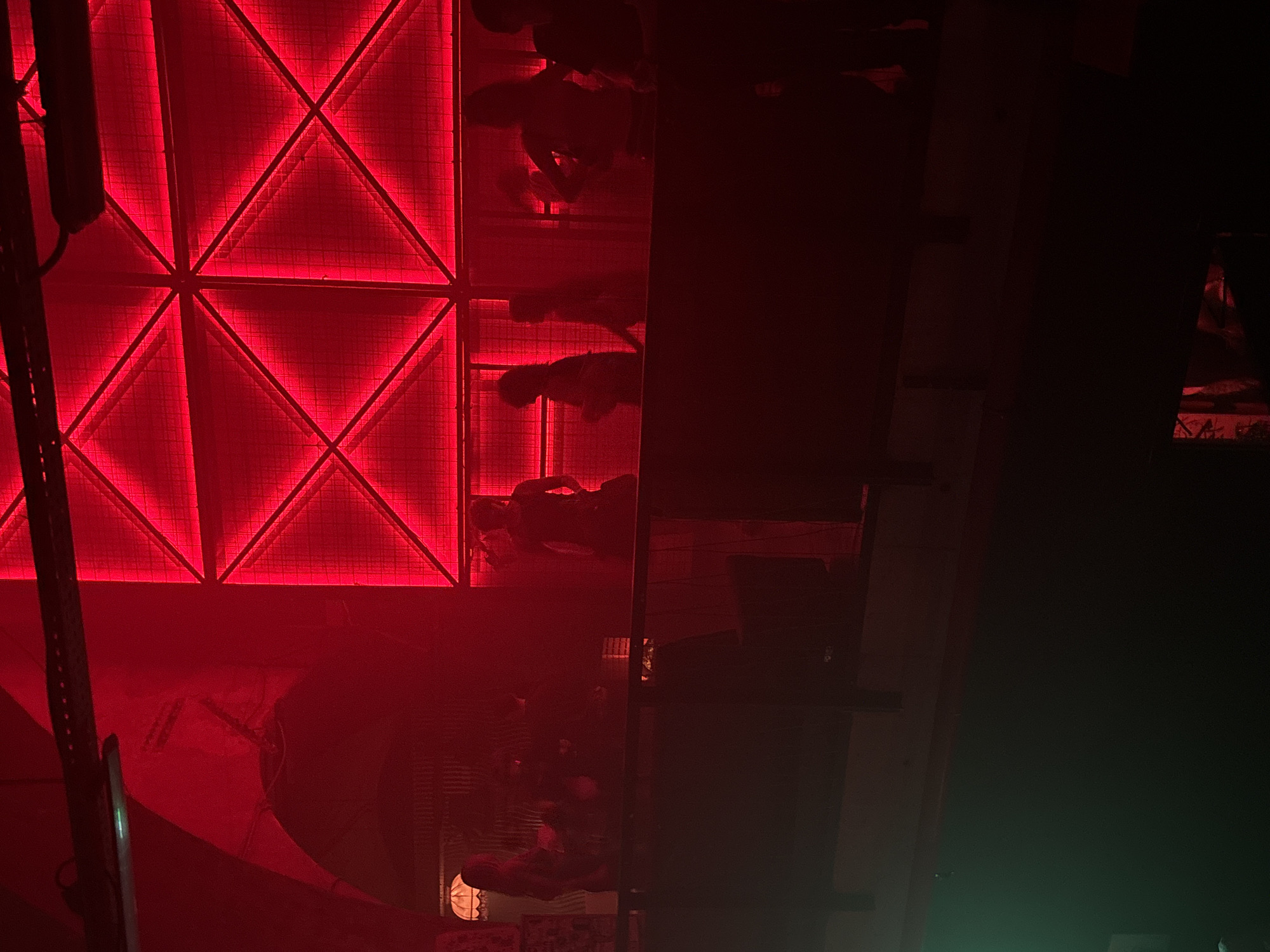 The image depicts an interior scene, likely an entertainment venue such as a nightclub or bar, illuminated predominantly by deep red light. The location is in the city of Unknown, Unknown.
In the background, on the right side, a large wall feature is visible, composed of a metal grid or mesh behind which bright red linear lights are arranged. These lights form a pattern of interconnected triangles and squares, creating a striking geometric illumination across the wall. To the left of this feature, an arched architectural element, possibly part of a ceiling or alcove, is washed in the same red light, suggesting an older building structure. Overhead, dark structural elements, possibly beams or equipment rails, span across the upper portion of the frame.
A dark, solid barrier or railing extends horizontally across the lower mid-ground, separating the viewer's perspective from the area beyond. Behind this barrier, approximately a dozen individuals are visible, mostly silhouetted against the intense red lighting. Their forms are indistinct, but they appear to be standing and congregating, some facing an unseen area to the right, others seemingly interacting within their groups. No specific actions like dancing or clear conversations are discernible due to the low light and silhouetting.
In the mid-left portion of the image, behind the barrier, a distinct lampshade with a scalloped or frilled edge is partially visible, emitting a softer, yellowish-white light, offering a contrast to the dominant red. Below the railing, in the lower left corner, a faint spill of green or cyan light is present. The overall environment suggests an active social gathering in a dimly lit, artificially illuminated setting designed for atmosphere.