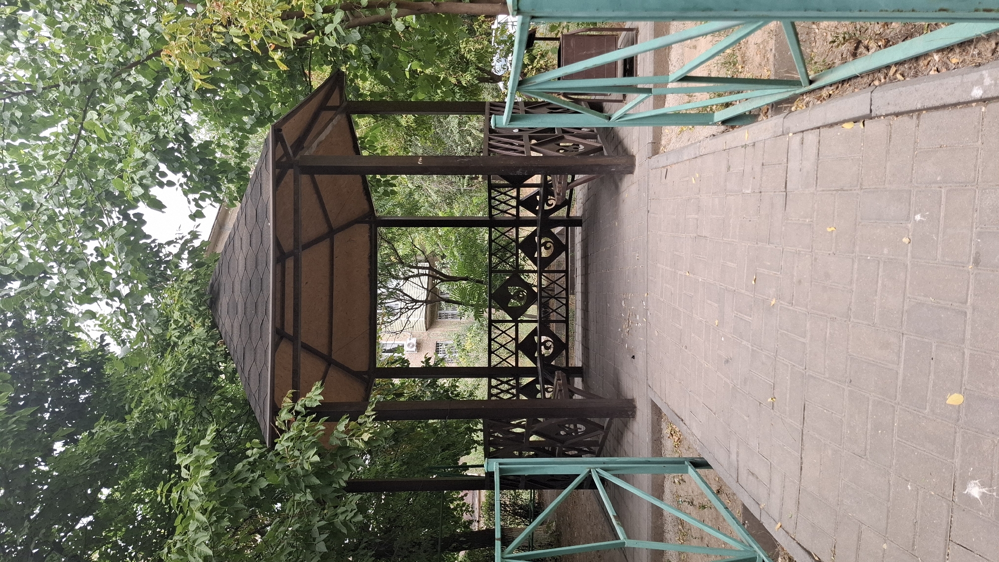 The image shows an outdoor gazebo with a dark, hexagonal roof supported by dark wooden posts. The gazebo has intricately patterned wooden railings and built-in benches. A paved walkway, made of rectangular grey bricks, leads directly to the entrance of the gazebo. On either side of the walkway, there are light teal metal fences. Beyond the gazebo, a multi-story building with visible windows and an air conditioning unit can be seen, surrounded by lush green trees. The ground is covered with fallen leaves, indicating an autumn season. The scene appears to be in a public park or residential area within the city of Shymkent, Kazakhstan, a common feature in such urban landscapes.