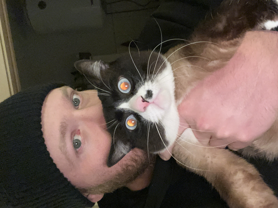 The image captures a close-up, vertically oriented portrait of a person holding a cat. The perspective suggests a selfie or a photo taken at an unusual angle, with the subjects appearing tilted to the side.
On the left side of the frame, a person with light skin, a beard, and what appears to be a dark beanie or cap is visible. His blue or green eyes are open and looking directly at the camera. He is wearing a dark top, possibly a black shirt or jacket.
Dominating the center and right side of the image is a cat, held by the person's hands. The cat's head is prominently featured, with its distinctive black and white markings. The cat has a black face with a white muzzle and a pink nose. Its eyes are a striking feature, glowing intensely with an orange-yellow hue, characteristic of the tapetum lucidum effect seen in flash photography. Its pupils are clearly visible. Long, white whiskers radiate from its muzzle. The cat's body appears to be a lighter, tan or cream color, contrasting with its darker head markings, suggesting it might be a bicolor or perhaps a Siamese-patterned cat. One of the person's hands is visible holding the cat around its body, with the fingers wrapped around its chest/belly area.
Both the person and the cat are looking towards the camera, suggesting they are posing for the photo. The background is dark and out of focus, providing little detail but indicating an indoor setting, possibly at night or in a dimly lit room. There are no visible texts or specific objects in the background to pinpoint the exact location or time. The overall impression is one of a warm, affectionate moment between a pet owner and their cat.