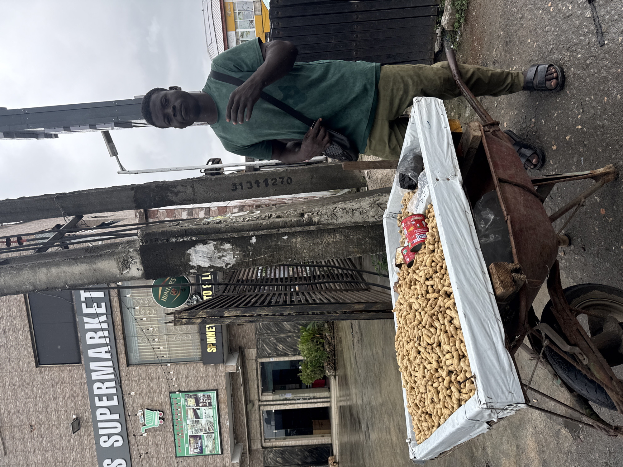 In the city of Ado, Nigeria, a young Black man stands next to a wheelbarrow filled with peanuts. He is wearing a green t-shirt, olive green pants, and black sandals. A black bag with a textured strap is slung across his body. The wheelbarrow is old and rusted, with a white tarp lining its basin. Two red cans are visible among the peanuts. Behind the man and the wheelbarrow is a building with "S SUPERMARKET" visible on its facade. A concrete pillar and a metal gate are also present in the scene. The ground is wet, suggesting recent rain.