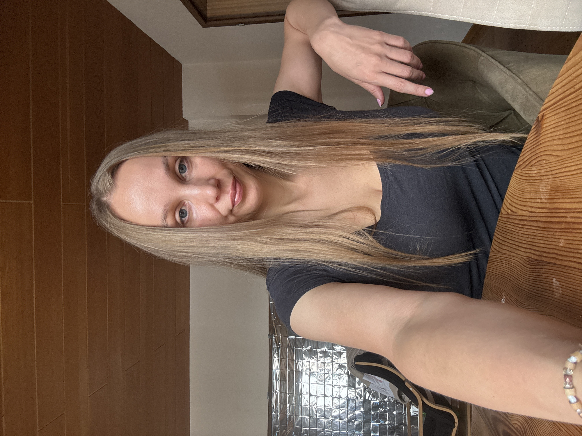 A woman with long blonde hair is taking a selfie in a room. She is wearing a dark gray t-shirt and is seated at a wooden table. Her right arm is bent and resting on the back of a chair, and her left arm is extended towards the camera. Her fingernails are painted pink. In the background, there is a metallic, tiled wall and a wooden ceiling. The scene appears to be indoors. There is no visible indication of the specific location within Überherrn, Germany.