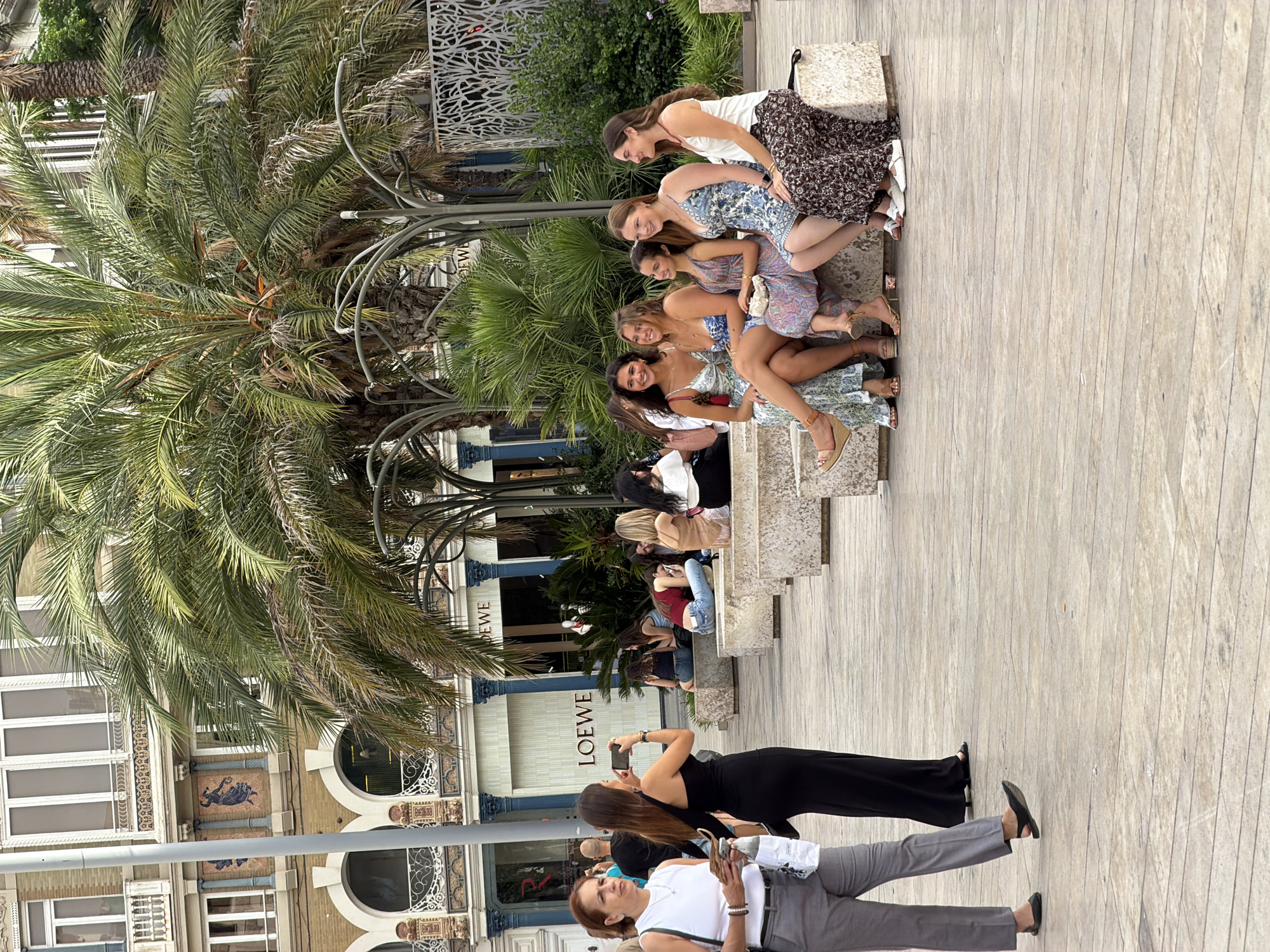 A medium shot captures an outdoor urban scene in front of a building with arched windows and palm trees. Several people are present. In the foreground, a woman in a white tank top and grey pants walks to the left, holding a phone and a white bag. Next to her, a woman in a black one-shoulder dress walks to the left, holding a phone up to take a picture. Behind them, a group of six young women sits on stone benches, appearing to be socializing and looking in various directions. The building in the background has ornate architectural details and signs that read "LOEWE". The ground is paved with wooden planks. The scene is illuminated by natural daylight.