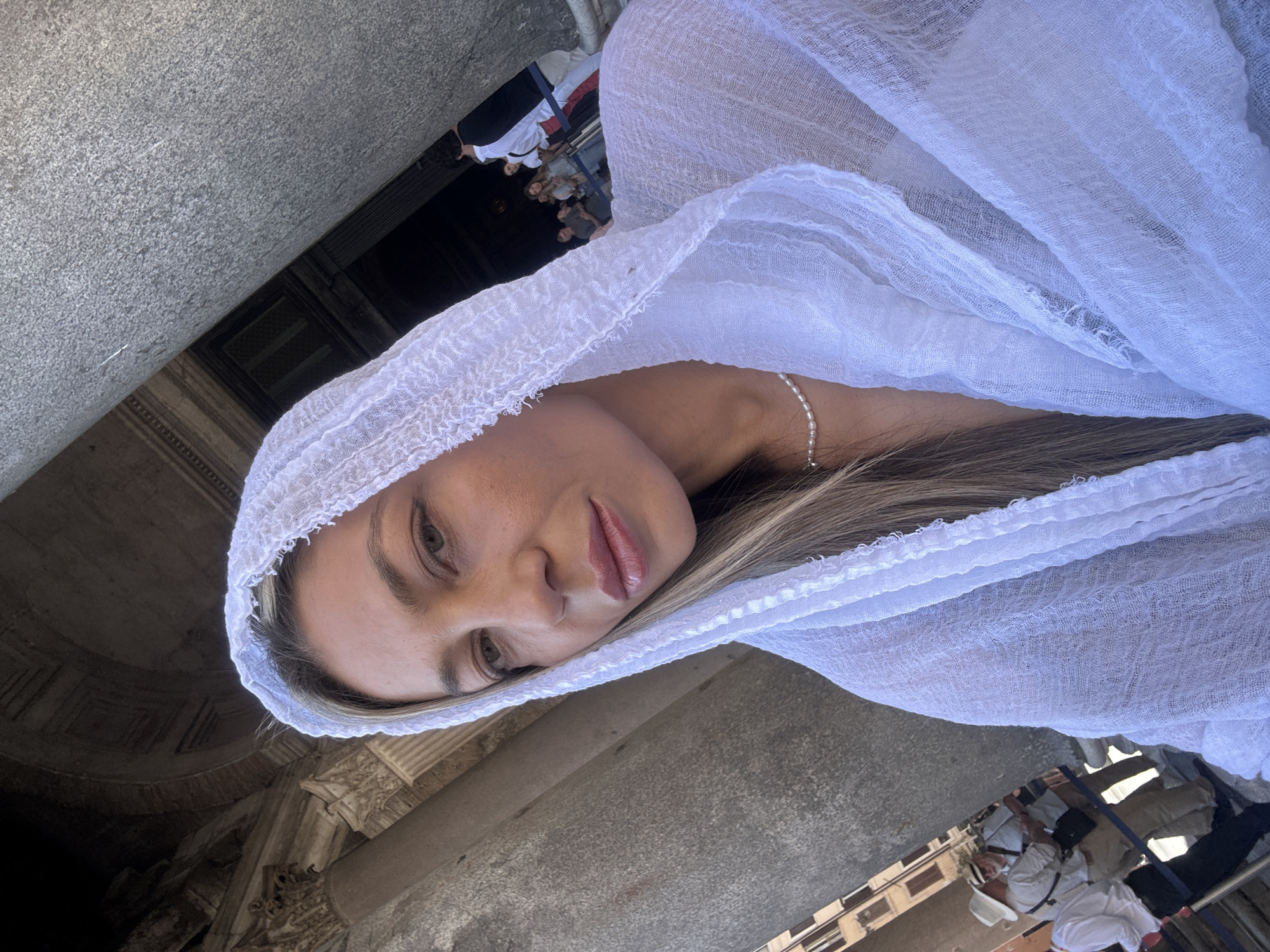 A medium shot photograph depicts a woman indoors, with a white fabric draped over her head and shoulders. The woman is wearing a delicate pearl necklace. A stone column is visible to her left, with other architectural elements present, including archways and moldings. In the background, the scene contains indications of other people and urban buildings. The specific location is the Pantheon in Rome.
