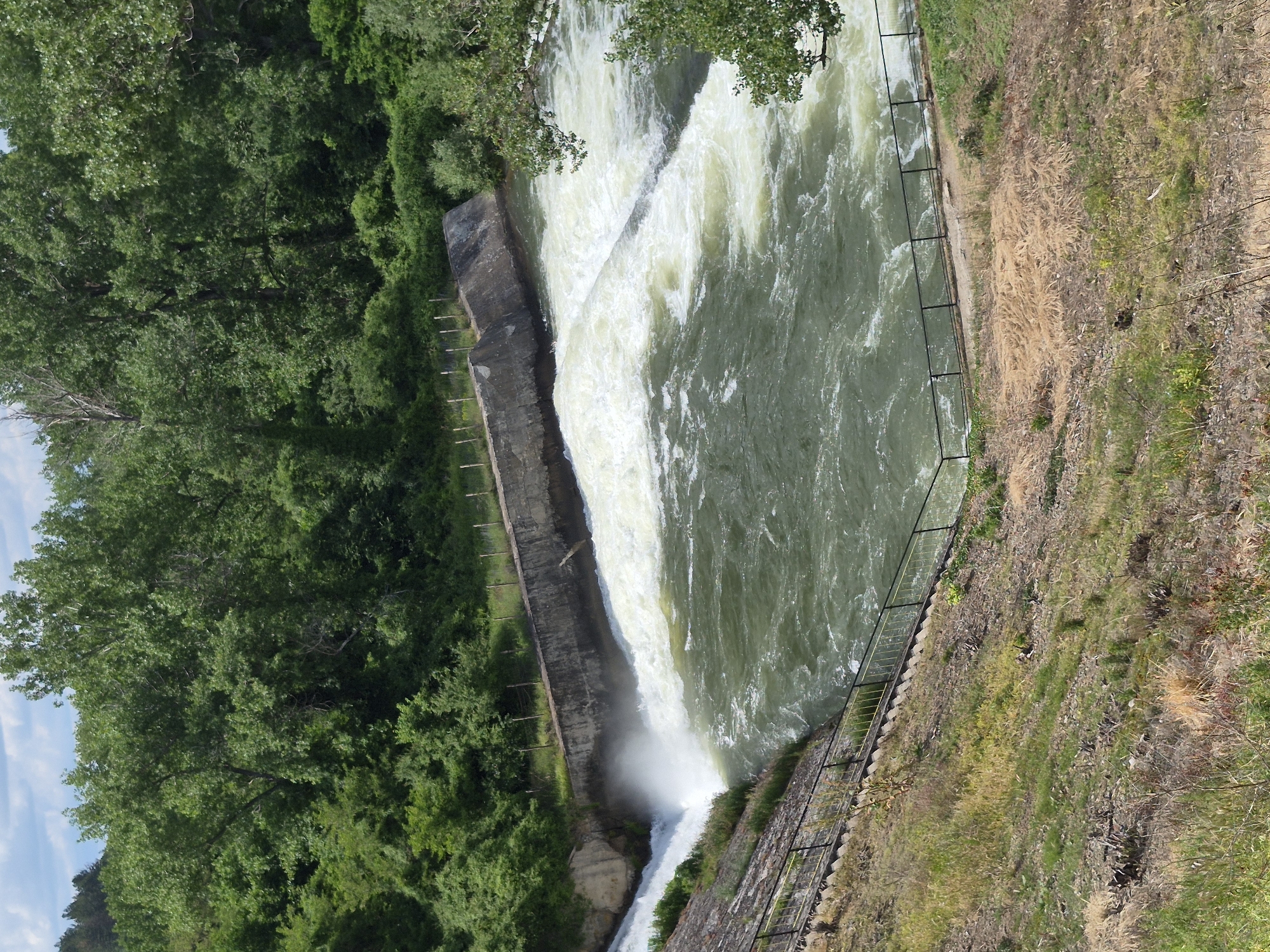 An aerial, high-angle, full shot captures a turbulent body of water flowing over a spillway. The water is a murky green, with white foam churned up from the spillway. Concrete structures flank the spillway, with the banks on either side covered in green foliage. A black metal fence lines the spillway and the embankment on the left side. The sky is partly cloudy, with hints of blue visible. The scene appears to be in a natural setting, possibly a dam or a natural waterfall. There are no people or animals visible in the image. The lighting is bright, suggesting daytime.