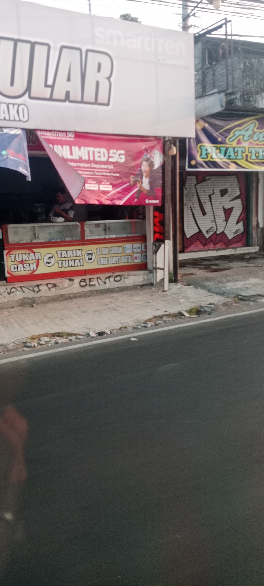 This is a street-level, slightly angled, medium-distance shot of a commercial establishment in Banyuraden, Indonesia. The main building features a large white banner with bold, silver lettering that reads "ULAR" in the foreground, and "smartfren" in smaller white text above it. Below this, a red banner advertises "UNLIMITED 5G" with a stylized image of a young person holding a phone and appearing excited. Further down, there are signs with red and yellow backgrounds indicating services like "TUKAR CASH" (Cash Exchange) and "TARIK TUNAI" (Cash Withdrawal), along with icons for financial transactions. The storefront itself appears to be a small shop with glass display cases. Graffiti in black spray paint is visible on the concrete wall and sidewalk in front of the establishment, with words like "HANIP" and "BENTO-" clearly legible. To the right of the main building, there is a shuttered storefront with graffiti in white and red spray paint, forming a stylized design. A dark road occupies the lower portion of the image, with a white line marking its edge. The overall impression is of a busy commercial street with various services and advertising.