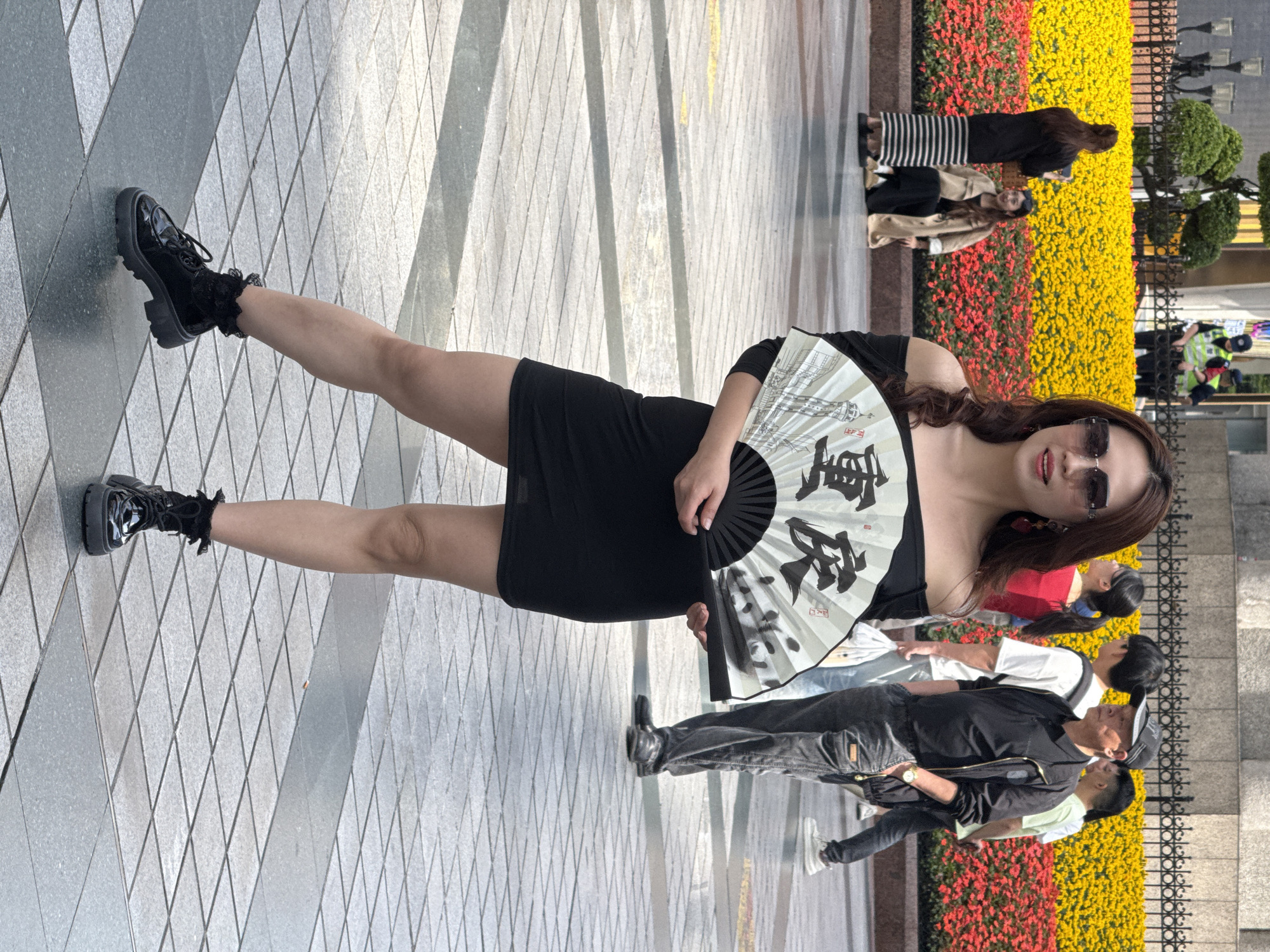 An outdoor, full-length shot shows a woman in a black dress posing on a tiled pedestrian walkway. She wears chunky black boots with frilly black socks and dark sunglasses, holding a white folding fan adorned with black Chinese calligraphy and an architectural illustration. The fan prominently displays the characters "重庆" (Chóngqìng). Her dark hair is styled around her shoulders.
In the background, a vibrant strip of red and yellow flowers creates a colorful border along the paved area, likely separating it from a street or another part of an urban landscape. Several other people are visible behind the woman, some walking casually, others standing near the flower beds. One person in the mid-ground wears a black jacket and jeans. Another individual in the deeper background wears a black and white striped dress. The scene appears to be taking place on a bright, sunny day, with clear lighting illuminating the ground and subjects. The setting suggests a lively urban environment, possibly a public square or shopping district in Yuzhong District, China, consistent with the text on the fan.