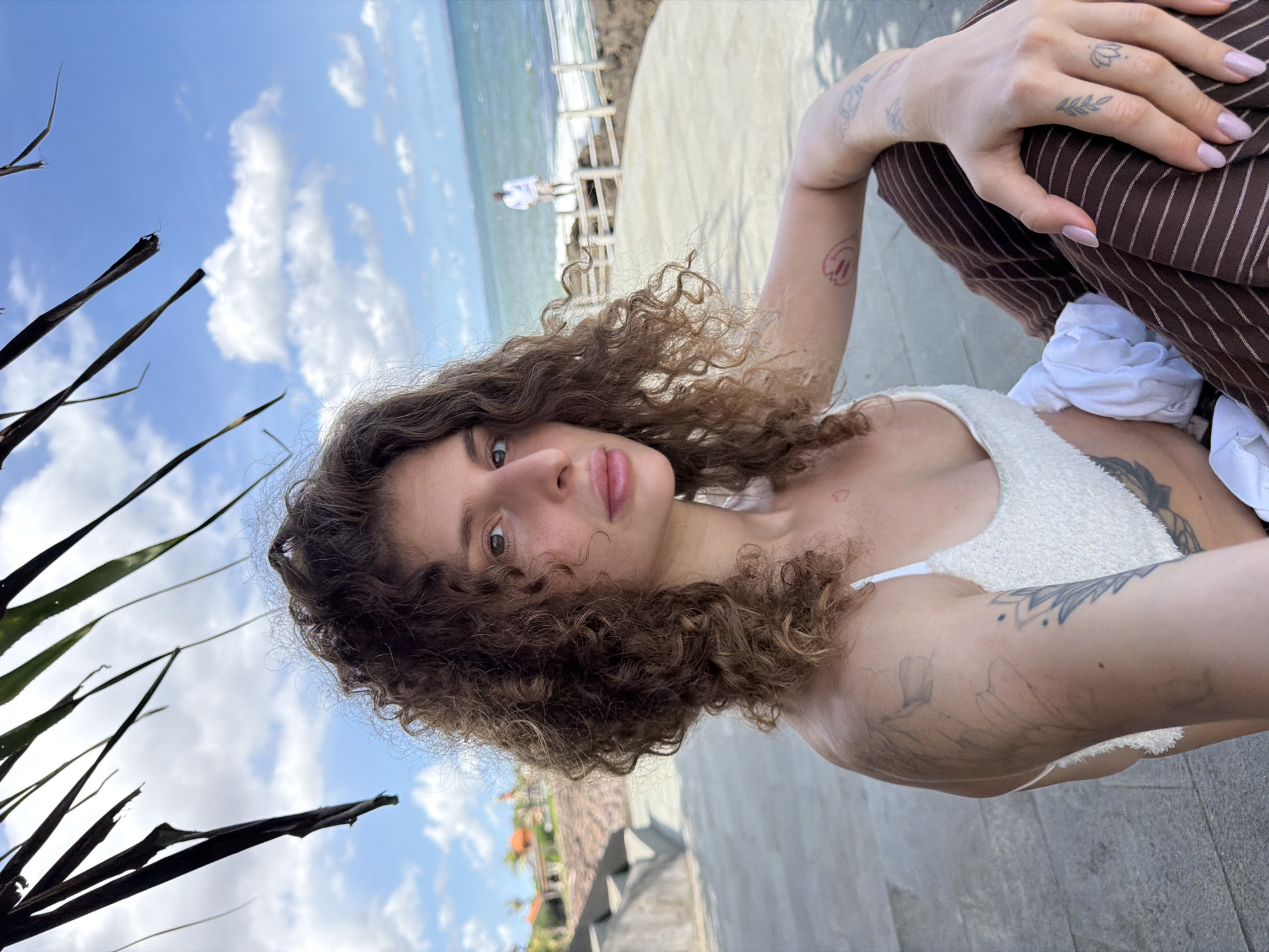 A woman with curly brown hair is photographed in Mengwi, Indonesia. She is wearing a white top and brown striped pants. She is sitting or reclining outdoors, near a coastline with a man standing in the background. She has multiple tattoos on her arms and hands. In the background is the sky with fluffy clouds. Part of a palm tree is visible on the left side.